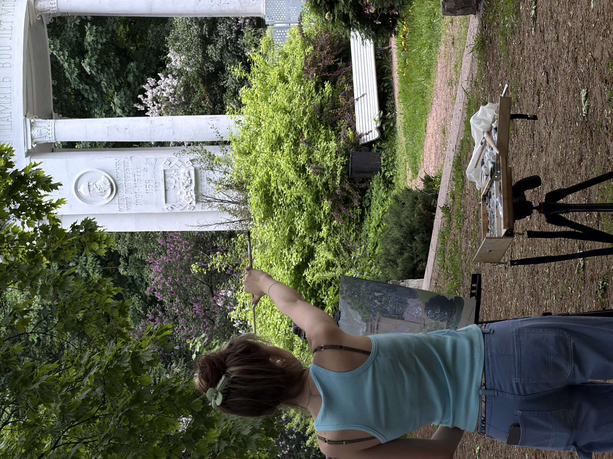 The image depicts a woman standing outdoors, painting on a canvas supported by a tripod. She is wearing a light blue tank top and blue pants. Her hair is tied up in a bun, with a green hair clip. She holds a paintbrush in her right hand. Behind her, there is greenery, including bushes and trees with purple and white flowers. To her left is a white monument with columns and inscribed text in Russian. In the background, there is a white bench. The painting setup includes a palette with paints and a container of brushes. The location appears to be in a park in Moscow, Russia, specifically near the "800 years of Moscow" monument.