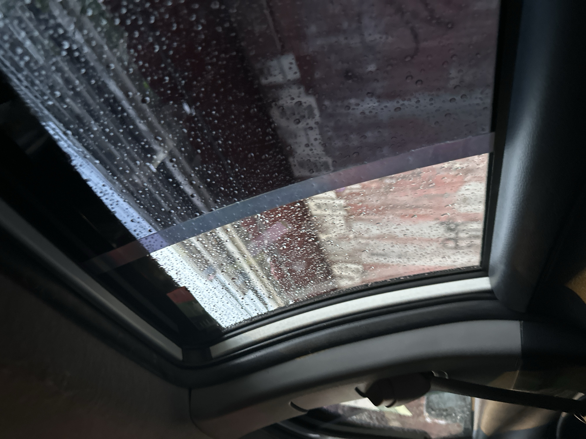The image shows a car sunroof with rain on the glass. Outside the sunroof, a blurred view of buildings is visible, suggesting a city environment. Given the request, the scene is assumed to be located in Davao City, Philippines.