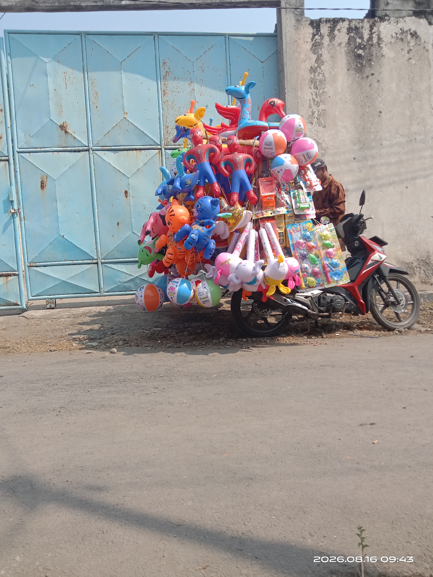 A vendor is parked on the side of a street in Banyuarang, Indonesia, with a motorcycle laden with a variety of colorful balloons and inflatable toys. The motorcycle is positioned in front of a large, weathered blue metal gate. The vendor, wearing a brown shirt, is seated on the motorcycle, facing away from the camera and towards the collection of goods. The balloons are shaped like animals, cartoon characters such as Spider-Man, and striped beach balls. Some of the toys are packaged, while others are deflated or partially inflated. The street is paved with asphalt and appears to be a typical urban road. The background shows a rough, textured concrete wall and a clear sky. The timestamp on the image indicates it was taken on August 16, 2026, at 09:43.
