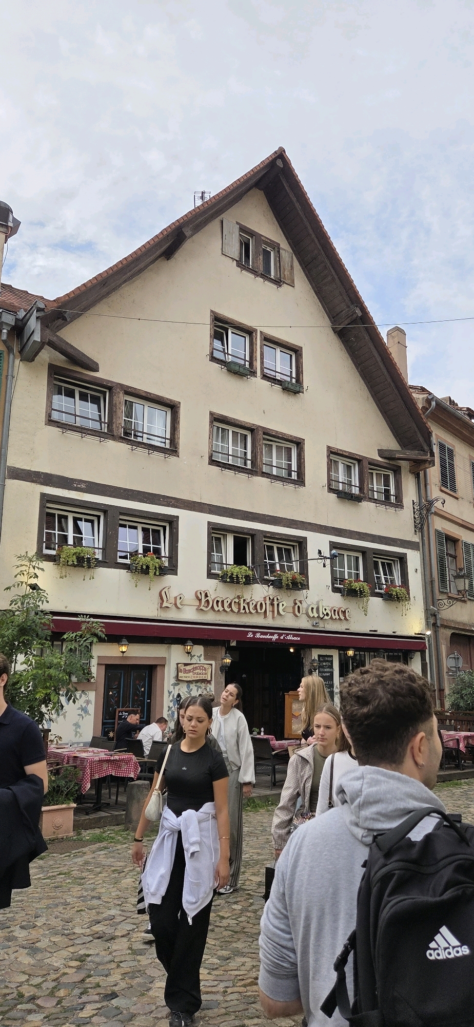 The image depicts a street scene in Strasbourg, France, featuring a traditional Alsatian building with a restaurant sign reading "Le Baeckeoffe d'Alsace". The building has a beige facade with dark wood trim and multiple windows with flower boxes. Several people are walking on a cobblestone street in front of the restaurant, which has outdoor seating with red and white checkered tablecloths. The sky is overcast. One woman in the foreground is wearing a black t-shirt and black pants, with a white sweatshirt tied around her waist. Another person in the foreground is wearing a grey hoodie and has a black backpack with an Adidas logo.
