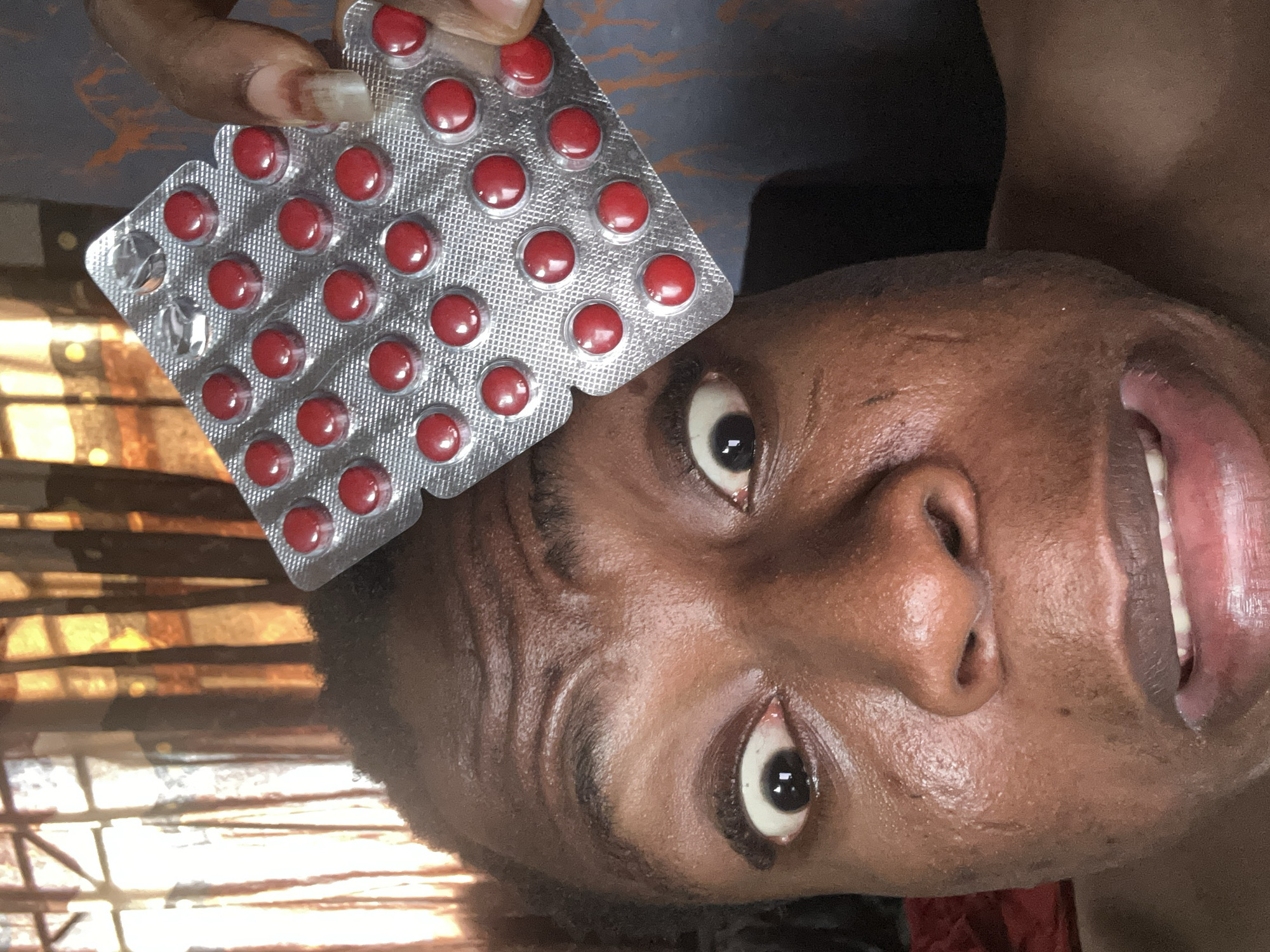 The image shows a dark-skinned individual holding a blister pack of red pills above their head. The blister pack contains 28 pills, with one pill missing. The person's face is visible, and they appear to be looking up at the camera. The background is blurred and indistinct. The location is unknown.
