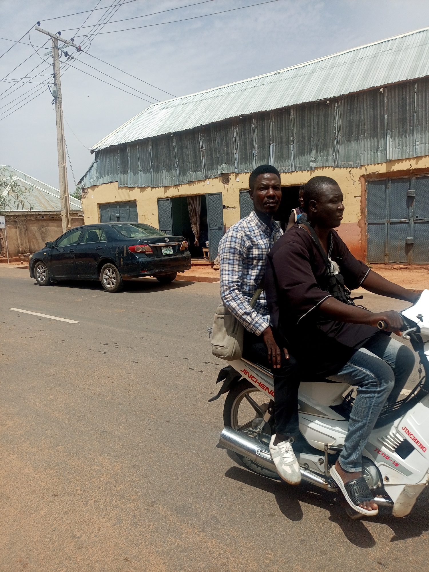 Two men are riding on a white Jincheng motorcycle on a street in Gombe, Nigeria. The driver is wearing a black shirt, blue jeans, and black sandals. The passenger is wearing a blue and white checkered shirt, dark pants, and white sneakers. He also has a tan-colored bag slung over his shoulder. A black sedan is parked on the side of the road. Behind the sedan and motorcycle is a tan-colored building with a silver, corrugated metal roof. A utility pole stands to the left of the building. The street is paved and dusty. The sky is bright and slightly overcast.