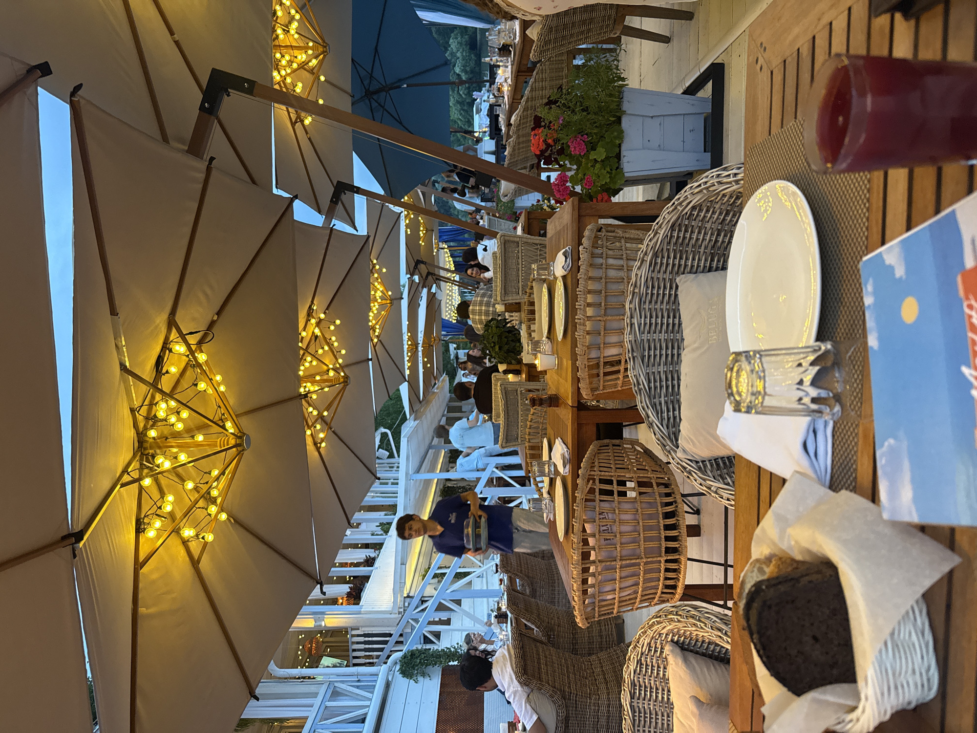 The image depicts an outdoor dining area at a restaurant in Zhukovka, Russia, likely during twilight. Numerous large beige umbrellas, adorned with strings of warm yellow lights, extend over several wooden tables set for dining. The tables are occupied by patrons and are furnished with wicker chairs, plates, glasses, and napkins. In the mid-ground, a waiter in a blue shirt and grey pants carries a tray of plates. A white building with a staircase and white railing is visible in the background, and a body of water can be seen further back, suggesting a waterfront location. The overall ambiance is relaxed and inviting, typical of an evening meal at a resort town.