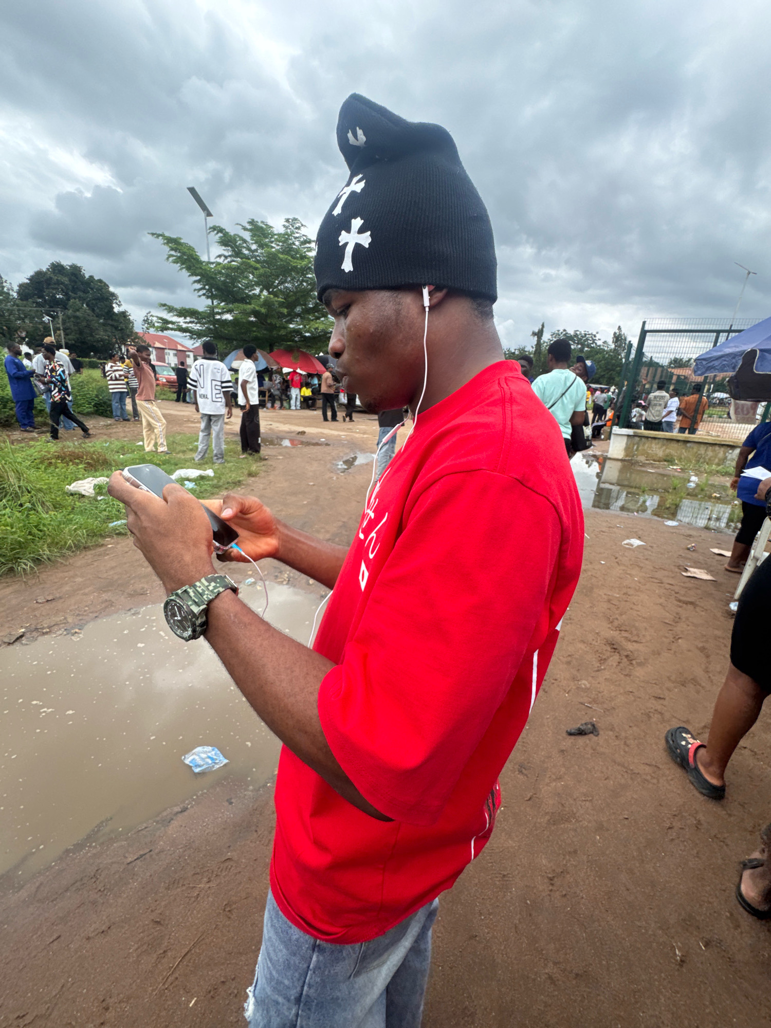 The image shows a young Black man standing in a dirt area with several puddles. He is wearing a black beanie with white crosses, a red t-shirt, and ripped blue jeans. He is holding a smartphone in his left hand and appears to be looking at it. A watch with a camouflage band is on his left wrist. In the background, there are other people, tents, and buildings. The sky is overcast. The scene appears to be a public gathering or market in Ihiagwa, Nigeria, possibly after a period of rain given the puddles and muddy ground.