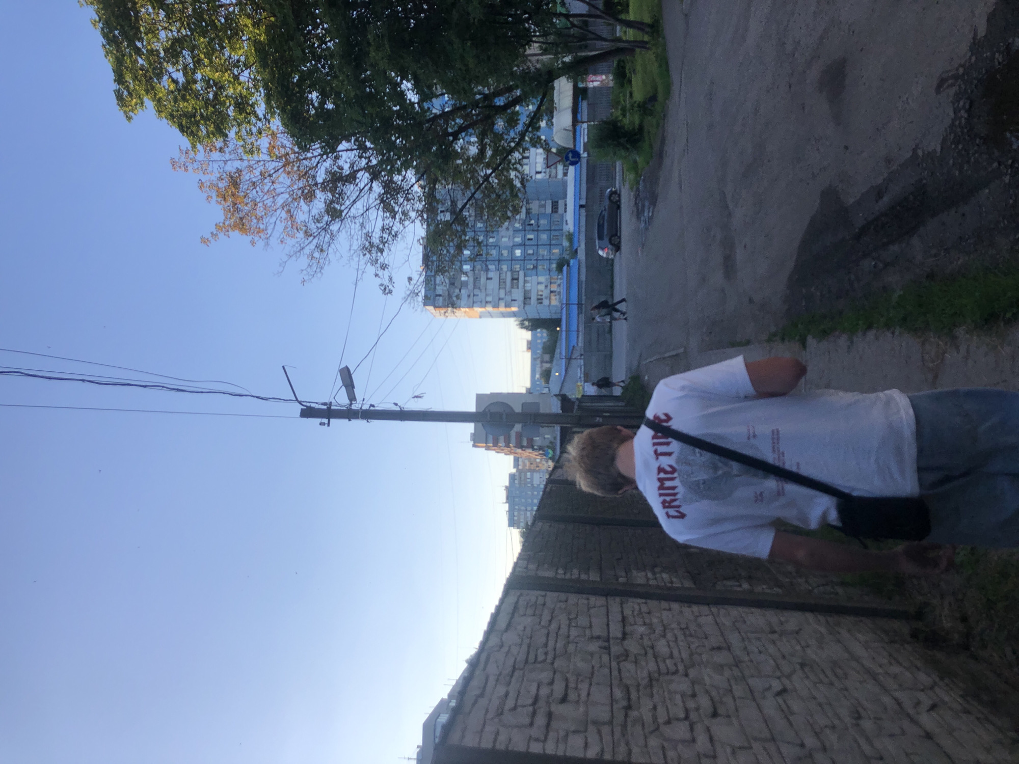 A person with blonde hair walks away from the camera down a dirt road, wearing a white t-shirt with red lettering that says "CRIME TIME" and a black messenger bag slung across their body. To the left is a tall, grey stone wall, and to the right is a road with some puddles. In the background, there are several multi-story apartment buildings, trees, and utility poles with wires. The sky is clear and blue. This scene is consistent with an urban environment in the city of Dnipro, Ukraine, likely in a residential area.
