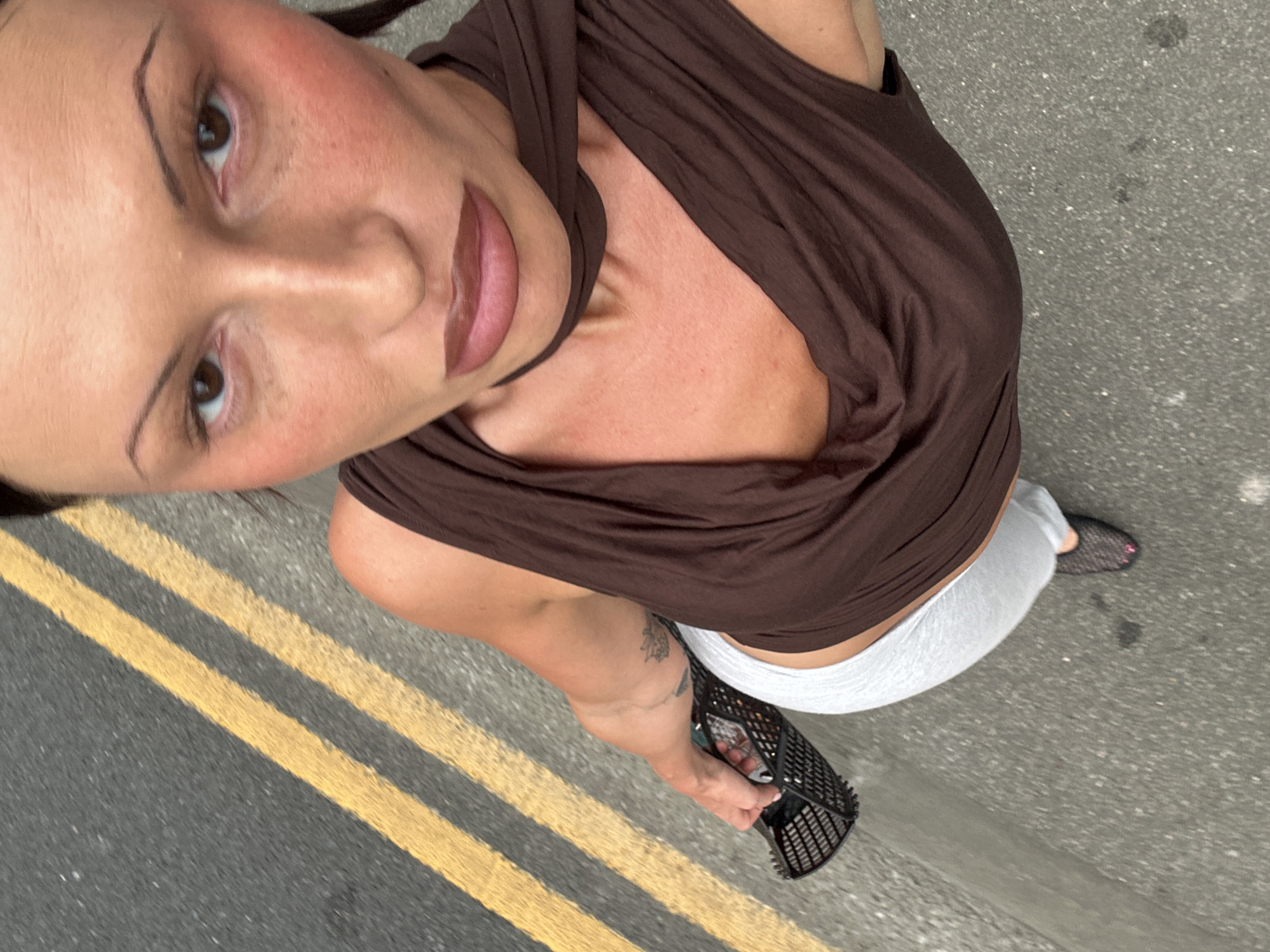 A woman is seen from a high-angle perspective standing on a road marked with double yellow lines. She is wearing a brown sleeveless top and white bottoms. In her left hand, she holds a black, basket-like object. The road surface is gray. Based on the yellow lines, it is likely in the city of Tunbridge Wells, United Kingdom.