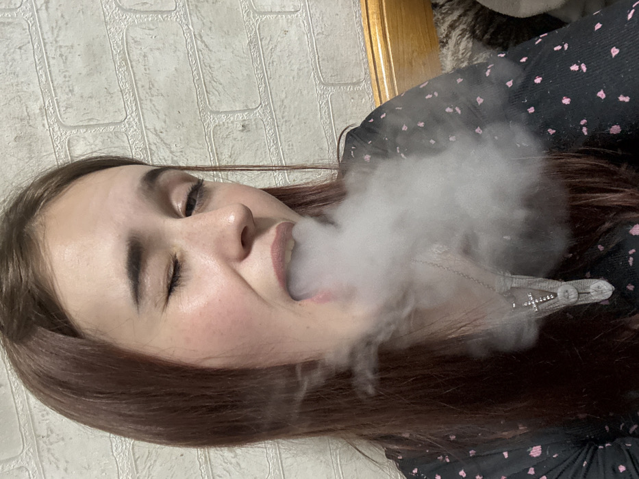 A young woman with auburn hair exhales a cloud of smoke. She is lying on what appears to be a bed, a wall behind her. She wears a dark blouse with pink floral accents. A silver necklace adorns her neck. She gazes towards the viewer with one eye partially closed.