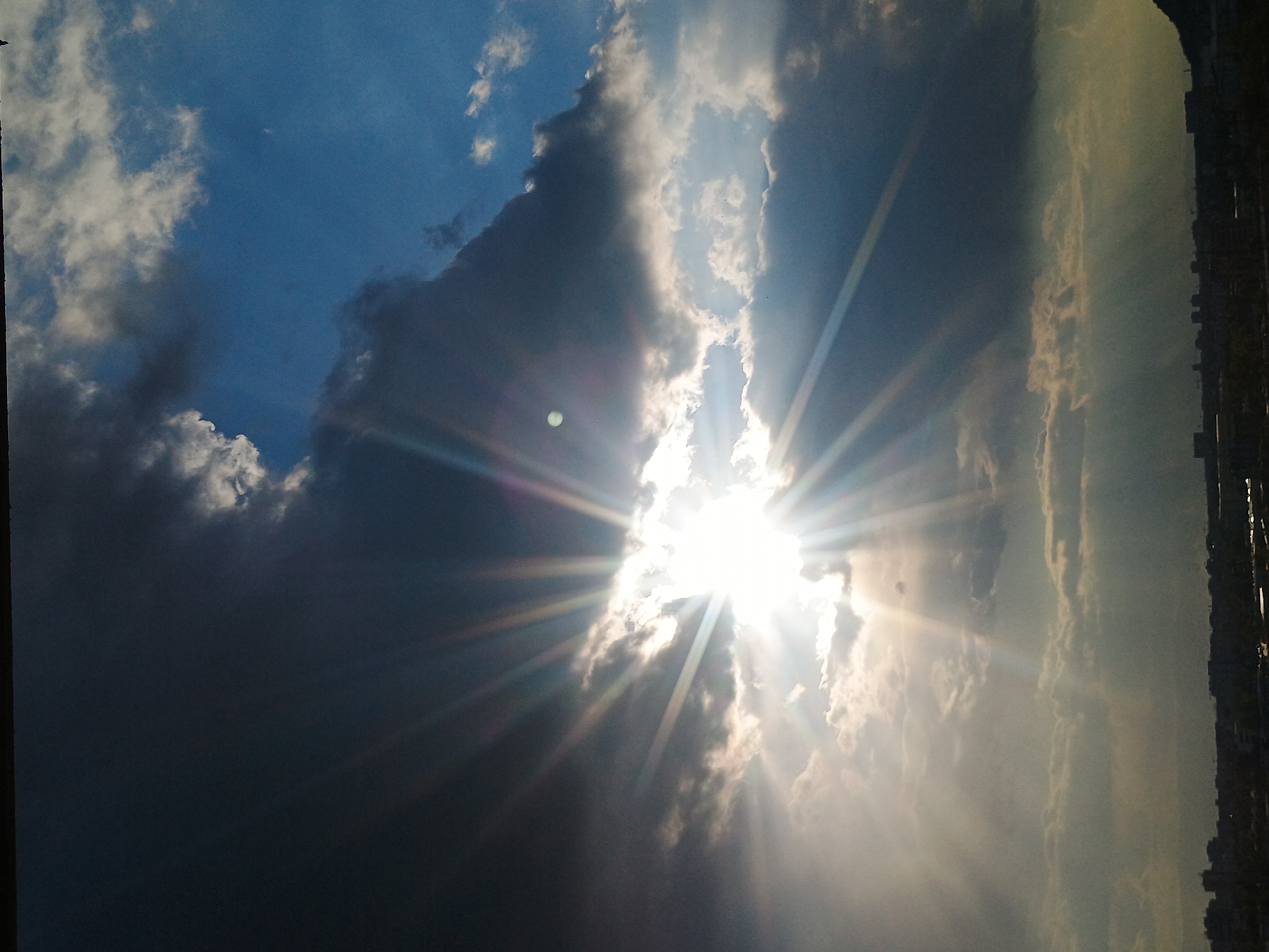The image shows a partially cloudy sky with the sun shining brightly through a gap in the clouds. The sun's rays are visible and spread out across the frame. Dark storm clouds are present on the left side of the image, contrasting with the bright sunlight and lighter clouds. A blurred cityscape is visible on the right side.
Without additional information, it's difficult to pinpoint the exact location within Plovdiv based solely on the image. The skyline offers little to no reference in terms of building shape or landmarks.