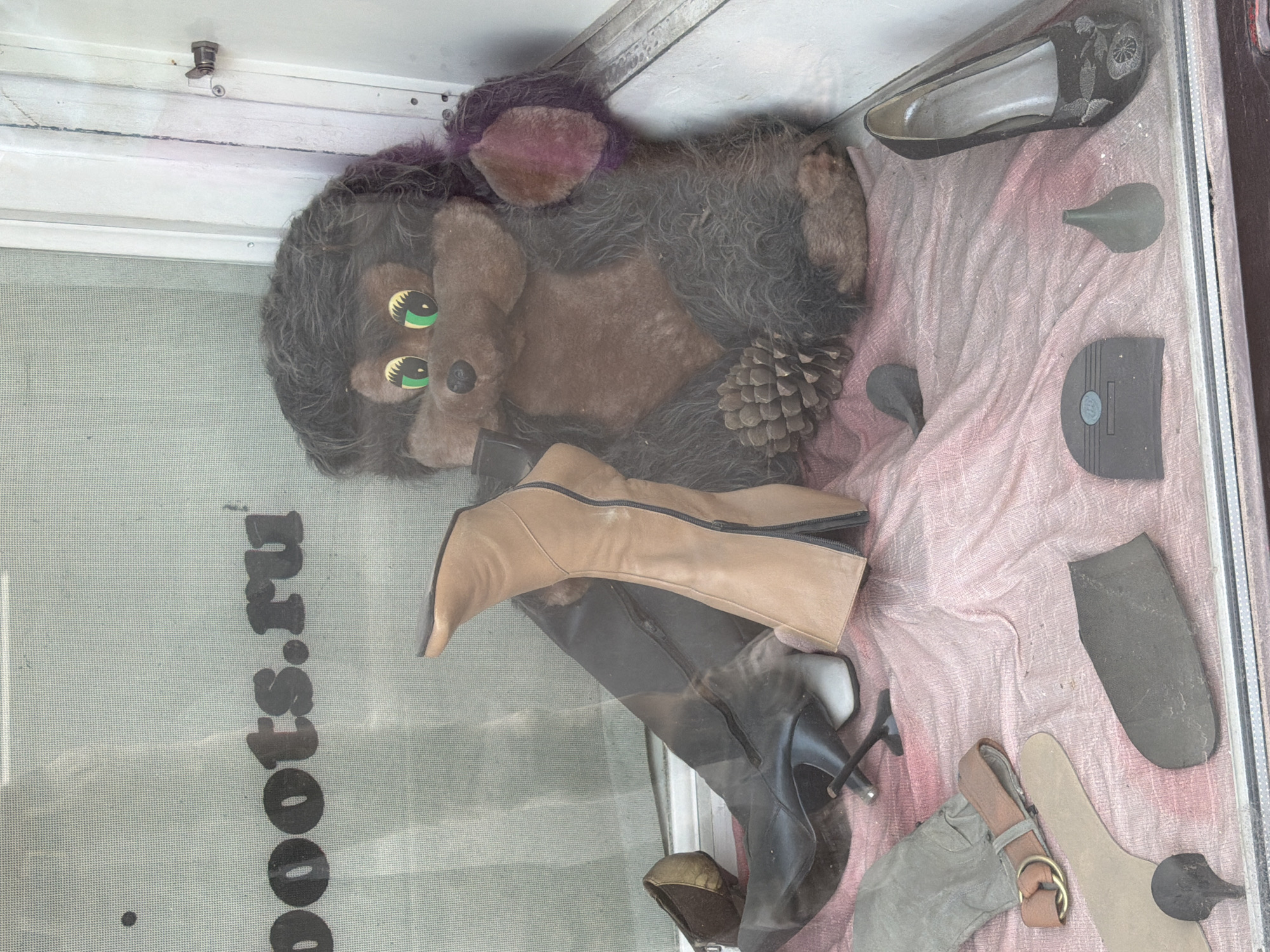 A plush toy animal with dark brown fur and bright green eyes is seated in a shop window display, propped up by a large pinecone. The toy is surrounded by various footwear and shoe-related items. To its left are several pairs of women's boots and shoes, including a tan calf-high boot with a zipper, a black knee-high boot with a stiletto heel, and a small brown shoe. Also present are shoe insoles and heel replacements. A pink fabric drapes the display. Behind the toy, text on the window reads "boots.ru", suggesting the location is a shoe store. The scene is viewed through glass, with reflections visible. The overall impression is a whimsical, slightly cluttered display in a retail setting.