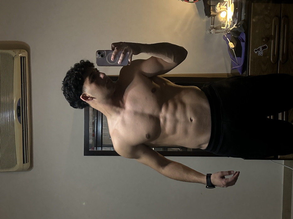 A shirtless man with curly black hair and dark pants takes a mirror selfie with his phone. He displays toned abs, chest, and arms. An air conditioning unit hangs on the wall to his left. Behind him is a framed mirror over what appears to be a window, with a dartboard and cabinet visible in the top right corner of the image. The lighting is dim and warm.