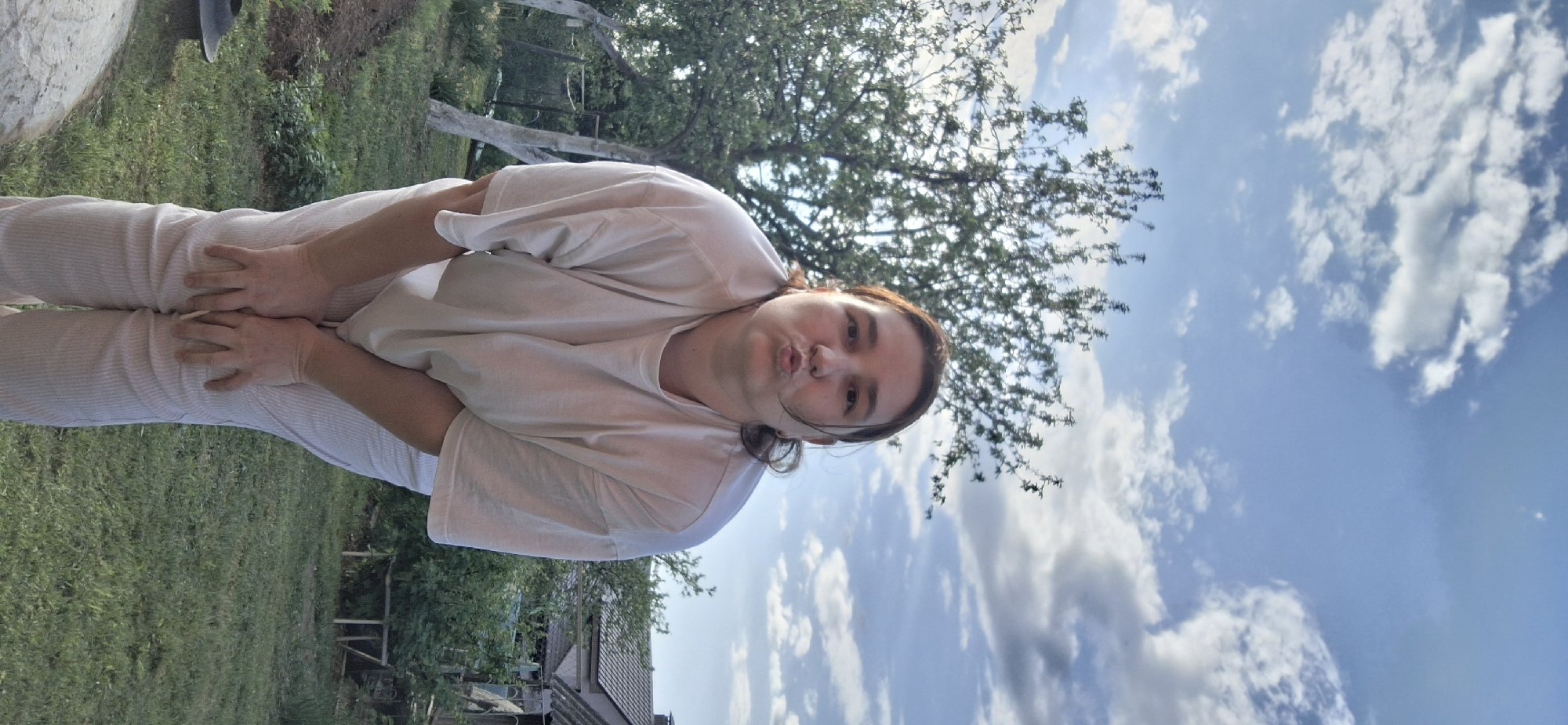 A medium shot of a person with light skin, brown hair, and wearing light colored clothing, standing outdoors. The person's hands are placed on their body, near the pelvic area. Behind the person, there is green grass and trees on the left, and a blue sky with clouds on the right. There is partial view of a house in the background. The location appears to be Chaikovsky, Russia.