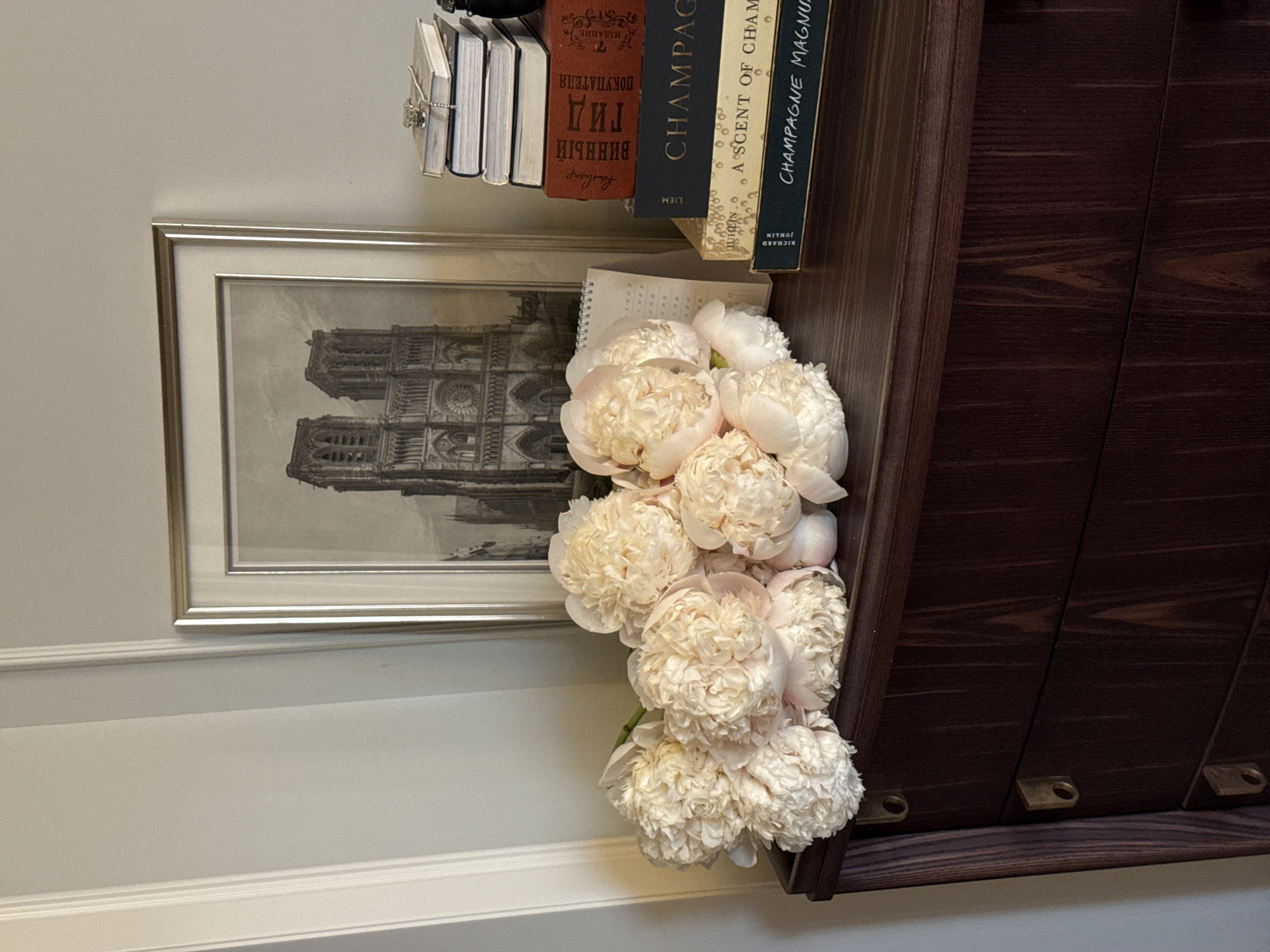 The image displays an interior still life featuring a bouquet of light-colored peonies, a framed picture, and a stack of books, all arranged on a dark wooden chest of drawers. On the left, a cluster of approximately ten peonies, predominantly cream or very pale pink, rests on the dresser surface. Behind them and slightly to the right, a silver-framed monochrome print or drawing of the Notre Dame Cathedral in Paris is visible. A small, white spiral-bound calendar or notepad stands upright between the peonies and the framed picture. To the right of the peonies and the calendar, a stack of books is arranged. The top of the stack consists of several small, light-colored notebooks, one of which has a silver chain or piece of jewelry placed on it. Below these are larger books: a red book with Cyrillic text "ВИННЫЙ ГИД ПОКУПАТЕЛЯ" (Wine Buyer's Guide), a dark blue book with gold text "CHAMPAGNE," a light brown book with "A SCENT OF CHAM" (likely "A Scent of Champagne"), and a dark green book with "CHAMPAGNE MAGNU" (likely "Champagne Magnum"). The furniture is a dark reddish-brown wooden chest of drawers or dresser with a curved front and three visible drawers, each featuring a small, brass-colored circular pull on the left side. The wood grain is distinctly visible on the furniture. The background wall is a light gray, with white decorative molding vertically positioned on the far left. The image contains no visual information or cues indicating that the location is in the city of Moscow, Russia. No individuals are present in the scene, and no actions are depicted.