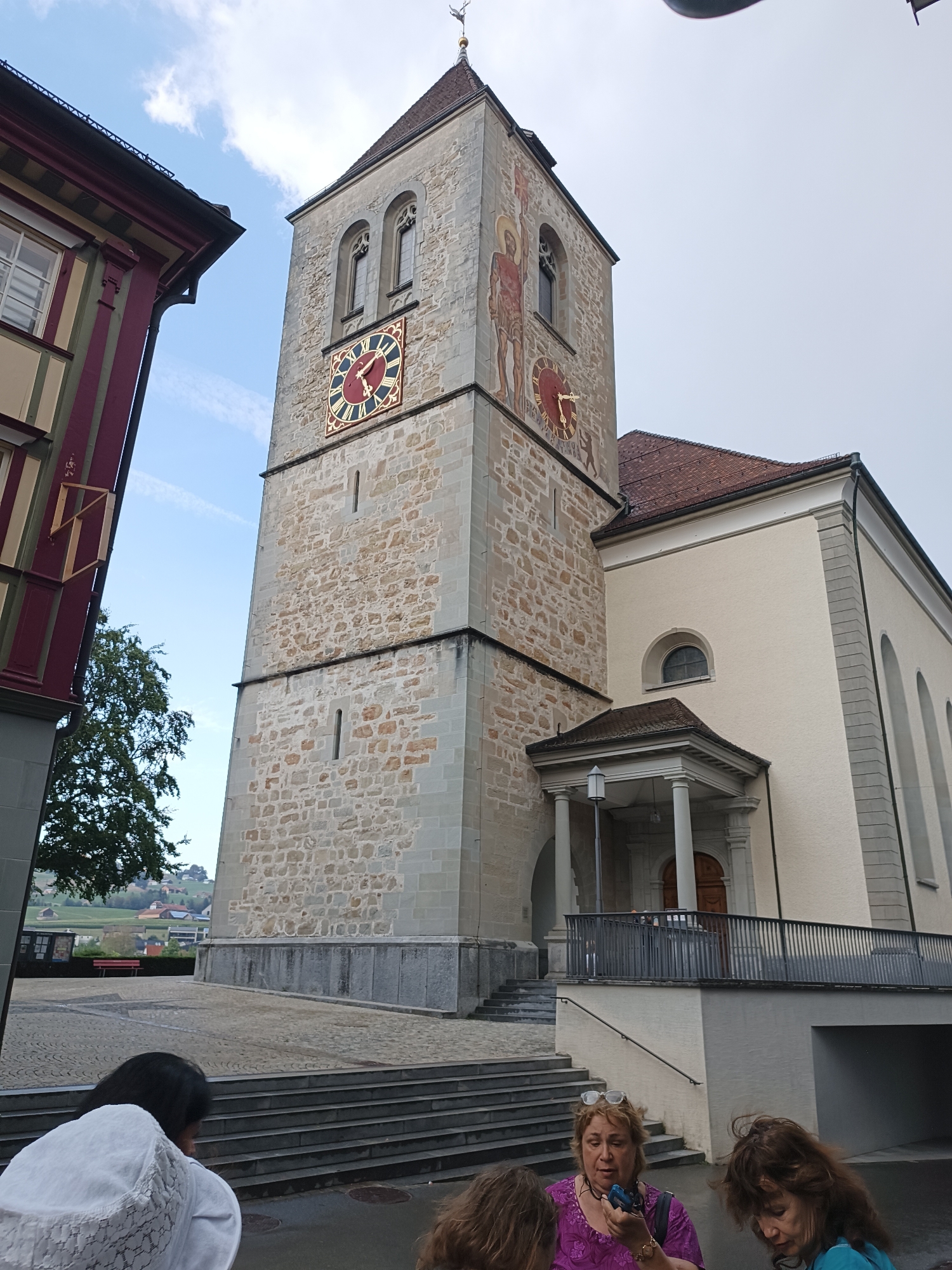 In Appenzell, Switzerland, a tall stone church tower with a prominent clock face dominates the scene. The clock, with Roman numerals and red and gold detailing, is situated on the tower's facade. To the left of the tower, a building with a dark red and cream striped facade is visible. In the foreground, three women are gathered. One woman, wearing a white hat, is seen from behind. Another woman, in a bright purple top, holds a phone and wears glasses. A third woman with brown hair is also present. The ground is paved with cobblestones, and there are stone steps leading up to the church entrance. In the background, green rolling hills and houses can be seen under a partly cloudy sky.