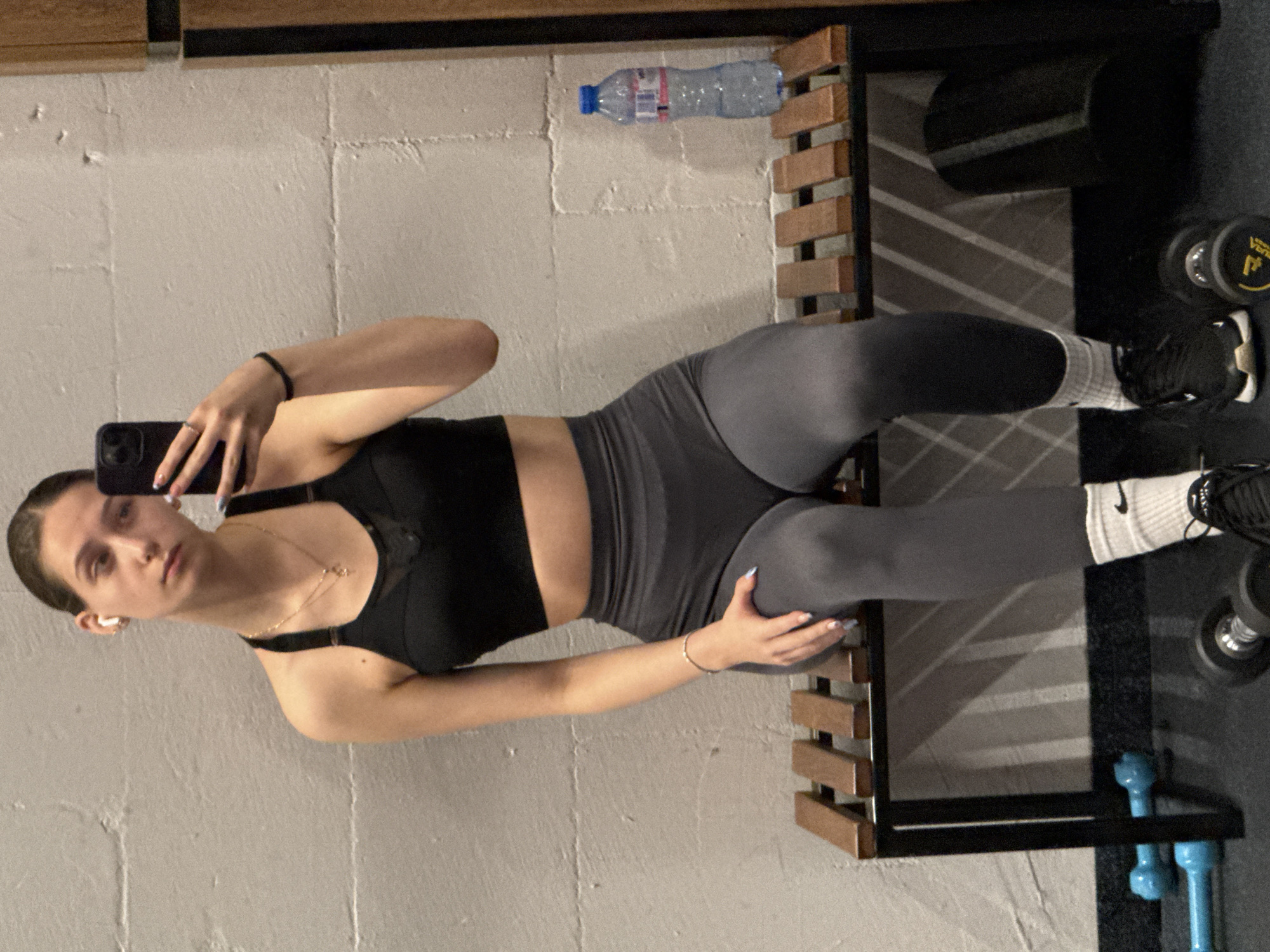 A woman takes a mirror selfie in a gym-like setting with weights and equipment. She wears a black sports bra, grey leggings, white socks with the Nike logo, and black sneakers. Her hair is pulled back, and she has a gold necklace and a black bracelet on her left wrist. She holds a smartphone in her left hand. The background includes a grey wall, a wooden and metal rack, a water bottle, and dumbbells. The location is unspecified but could be Sofia, Bulgaria.