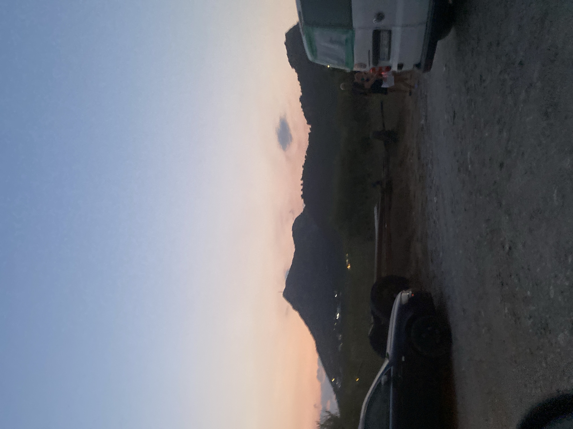 In Sutomore, Montenegro, at dusk, a scene unfolds with a dark-colored sedan parked on the left, facing right, with its front wheel turned slightly towards the viewer. Behind it, a pile of what appear to be large tires rests on the ground. Further to the right, a white van is parked, facing left, with its rear visible. Beside the van, two individuals, a male and a female, are standing, with the female holding what looks like a piece of paper. The background features silhouetted mountains against a sky displaying gradients of soft orange and pale blue, indicative of twilight. Scattered lights are visible on the mountainside, suggesting a settlement. The ground in the foreground is composed of gravel and dirt.