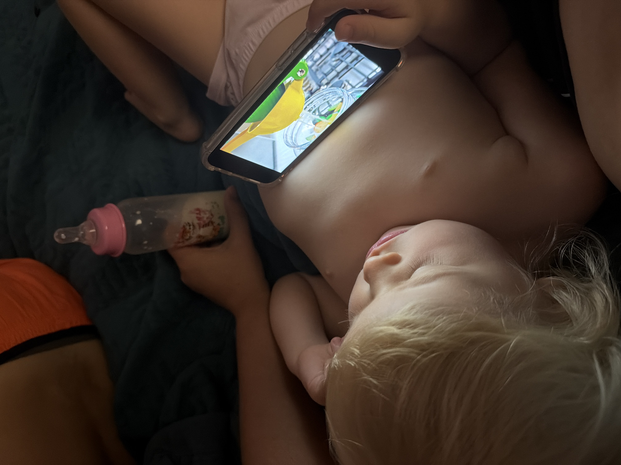 This is a close-up, eye-level, indoor photograph of a baby lying on their back, watching a mobile phone. The baby has blonde hair and fair skin, and is wearing pink bottoms. The phone is held in the baby's right hand and displays a cartoon of a yellow pitcher pouring liquid into jars. A baby bottle, partially filled with milk, is held in a person's left hand, positioned to the left of the baby. The background is dark and appears to be a bed with a dark green blanket. The overall lighting is dim, with the phone screen providing the primary light source. There is no information available about the specific location in Sochi, Russia.