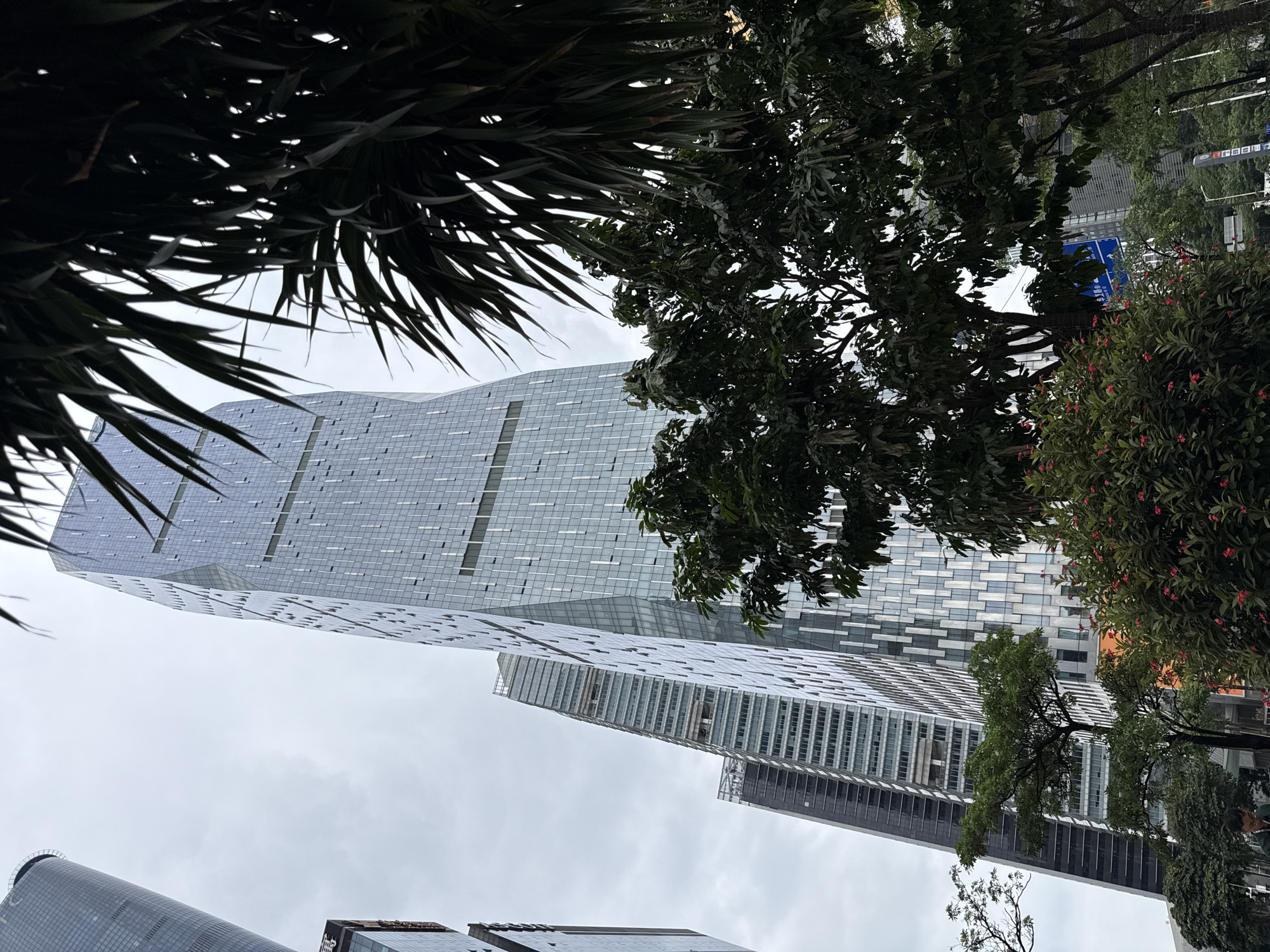 The image shows a street view in Tianhe District, China, dominated by modern high-rise buildings. In the foreground, lush green trees and bushes with small red flowers line the sidewalk. Beyond the foliage, several skyscrapers rise into a cloudy sky. The most prominent is a tall, slender building with a glass facade that twists as it ascends, reflecting the overcast sky. To its left, other tall buildings with similar glass exteriors are visible, some appearing more uniform in design and others with visible structural elements. The overall impression is one of urban development and a bustling cityscape.