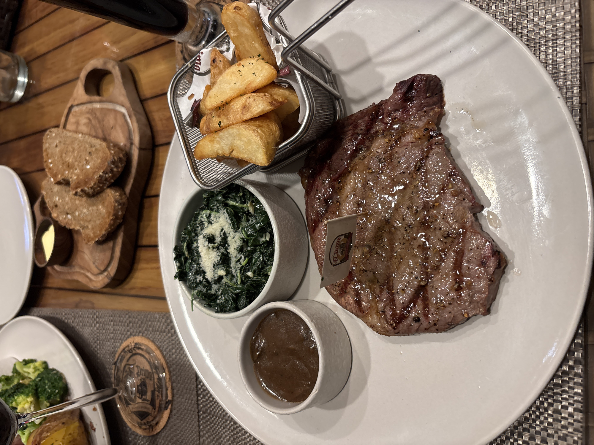 A top-down view shows a prepared steak dinner at El Cuccina restaurant, located in Kuta Selatan, Indonesia. The food is plated on a large, white round plate. It features a grilled steak, a small bowl of creamed spinach topped with cheese, a small bowl of brown sauce, and a wire basket of wedge-cut fries. A small card is sticking out of the steak. To the left, on a wooden board, there are three slices of bread and a small dish of butter. In the lower left corner, there is another white plate, partially visible, with broccoli and a baked potato. A glass of red wine is visible at the top of the image, near the wooden board. A glass with a coaster under it is present at the bottom left of the frame. The plate is set on a gray placemat. The wooden table surface is visible as well.
