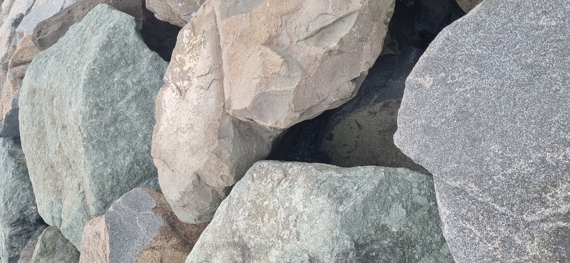The image displays a close-up view of a pile of large, irregularly shaped rocks. The rocks vary in color, with some appearing to be a light grey, speckled granite, while others are a dusty beige or a muted green-grey. The textures are rough and varied, with visible grains and some weathered surfaces. The rocks are densely packed together, creating shadows and crevices between them. Some of the rocks have a smooth, almost polished appearance in certain areas, likely due to erosion or water exposure. The lighting suggests an overcast day or shade, as there are no harsh shadows. The scene is devoid of any human presence or discernible activity. This is a photograph of a section of the Batumi Boulevard, specifically the area where the promenade is lined with large, decorative rocks, likely serving as a breakwater or an aesthetic element along the coastline of Batumi, Georgia.