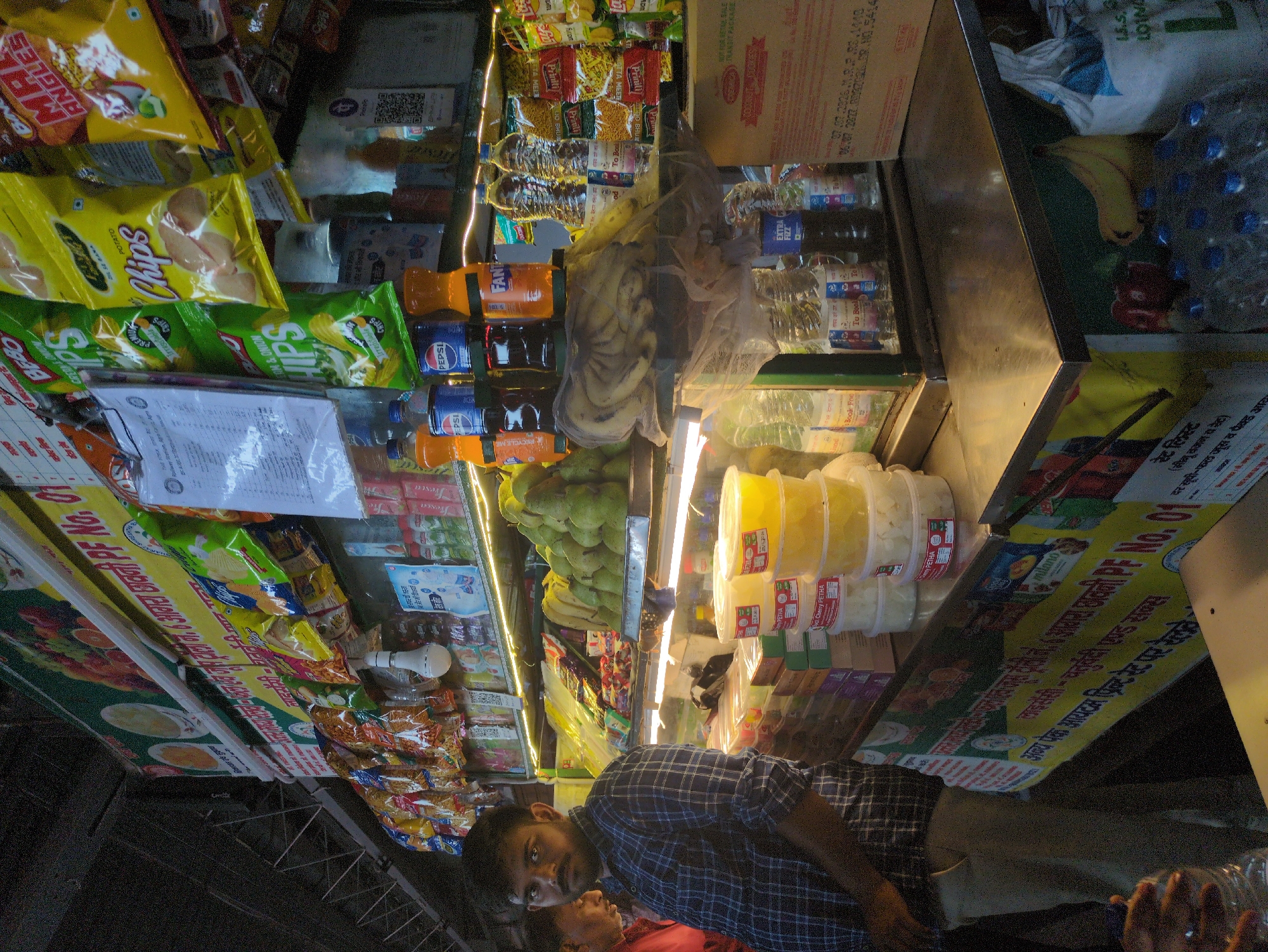 The image shows a roadside food stall in Agra, India. The stall is stocked with various packaged snacks, drinks, and fruits. A man in a checkered shirt stands in front of the stall, appearing to be the vendor or a customer. The stall has a prominent banner with text in Hindi, suggesting it is a licensed establishment. The background shows a variety of products arranged on shelves, including chips, biscuits, and bottled beverages. There are also fresh fruits like bananas and pears displayed. The overall scene depicts a busy marketplace environment typical of Indian cities.