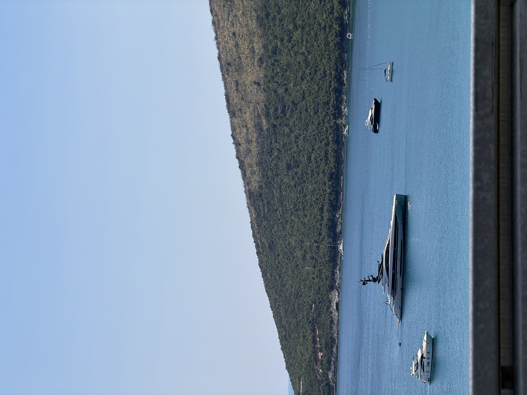 The image shows a bright, clear day with a wide expanse of turquoise water in the foreground. On the water, there are several boats. The largest is a luxurious white yacht with a sleek design and multiple decks, anchored in the middle of the frame. To its left, a smaller white motorboat is visible. Further out in the water, a dark-hulled motorboat is positioned to the right of the large yacht, and a white sailboat with its sail down is to the right of that. In the far distance, a verdant, tree-covered hill slopes down to the water's edge, forming a natural backdrop. The sky is a pale, cloudless blue. A portion of a grey railing is visible at the very bottom of the frame, suggesting the photo was taken from a viewpoint above the water. The location is identified as Radovići, Montenegro.