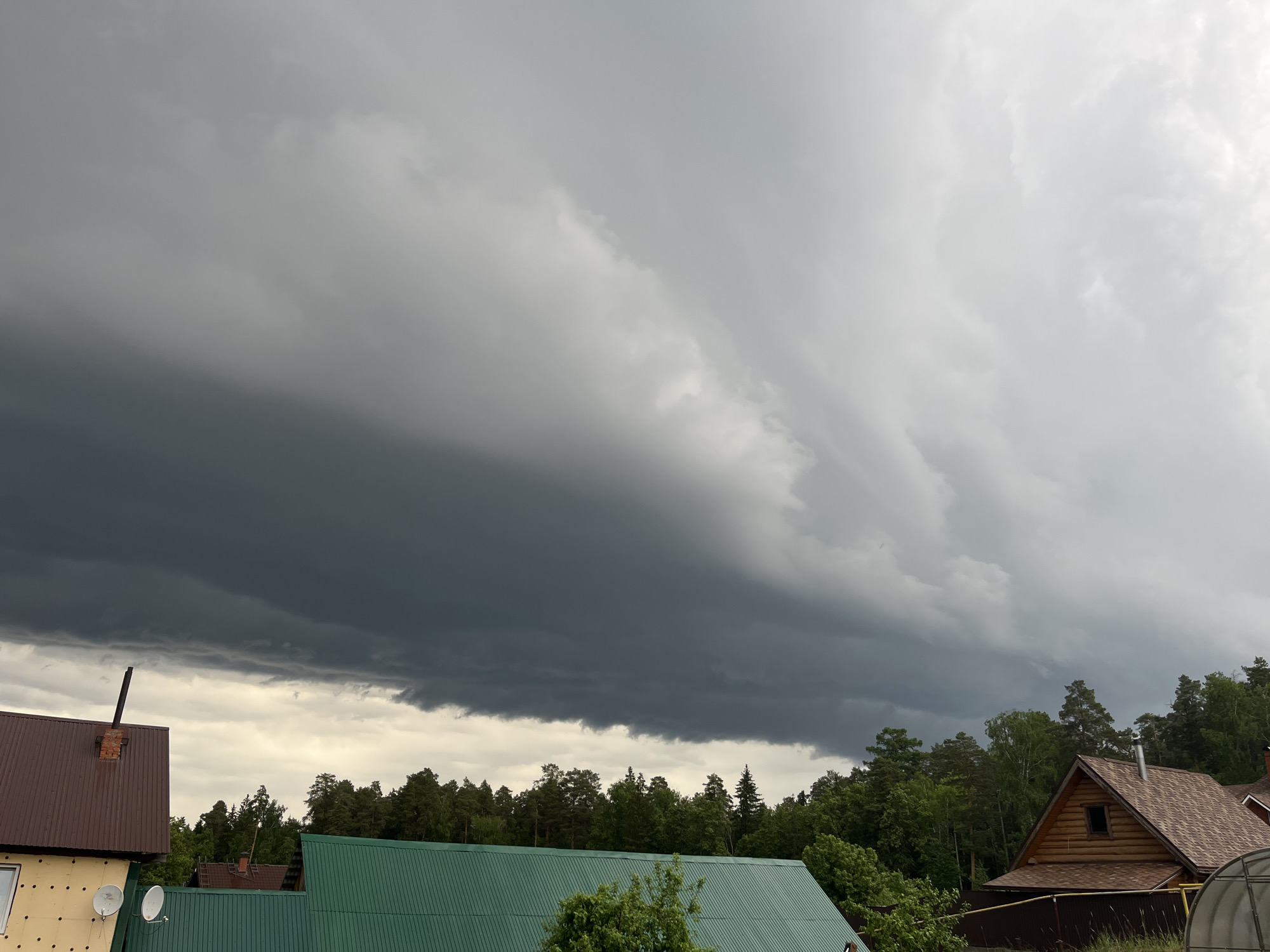 The image depicts a threatening storm cloud formation over a residential area in Chaikovsky, Russia. The dark, heavy cloud dominates the sky, contrasting with a lighter, overcast area. Below the clouds are rooftops of several houses with green and brown roofs, partially obscured by trees. A forest line is visible between the houses and the storm clouds. A satellite dish is mounted on a light yellow building. A brick chimney rises from the brown rooftop of a building on the left of the frame. The overall scene suggests an impending storm.
