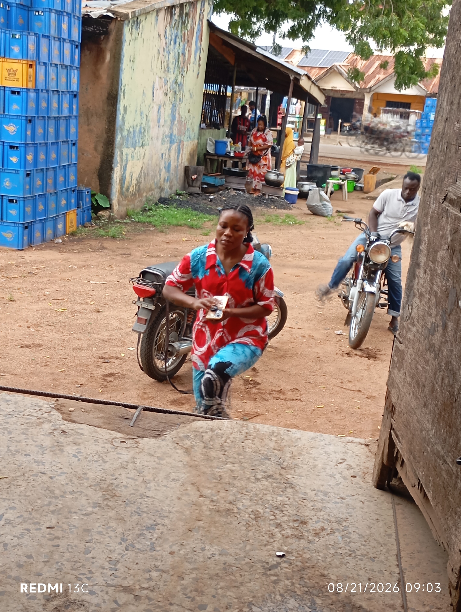 This is a photograph taken in Shendam, Nigeria, on August 21, 2026, at 09:03. The scene depicts a dusty outdoor market area. In the foreground, a woman in a red and blue patterned shirt and matching pants is kneeling next to a motorcycle. She is holding what appear to be a few bills in her hands. Behind her, a man wearing a light-colored polo shirt and blue jeans is riding a motorcycle. To the left of the scene, stacks of blue plastic crates are visible. In the background, there are several market stalls with corrugated metal roofs, some with people in them. Solar panels are visible on the roof of one building. The ground is covered in reddish-brown dirt and scattered debris.