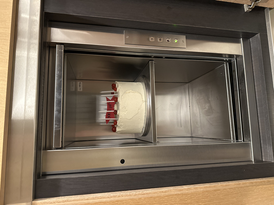 A white frosted cake with red decorations, including candles and a "Joe's 26th" banner, sits inside a stainless steel food elevator. The elevator is framed by dark brown wood, and a control panel is visible above the opening. The elevator's interior is brightly lit, creating a clean and sterile appearance. The surroundings suggest the elevator is located within a kitchen or dining area.