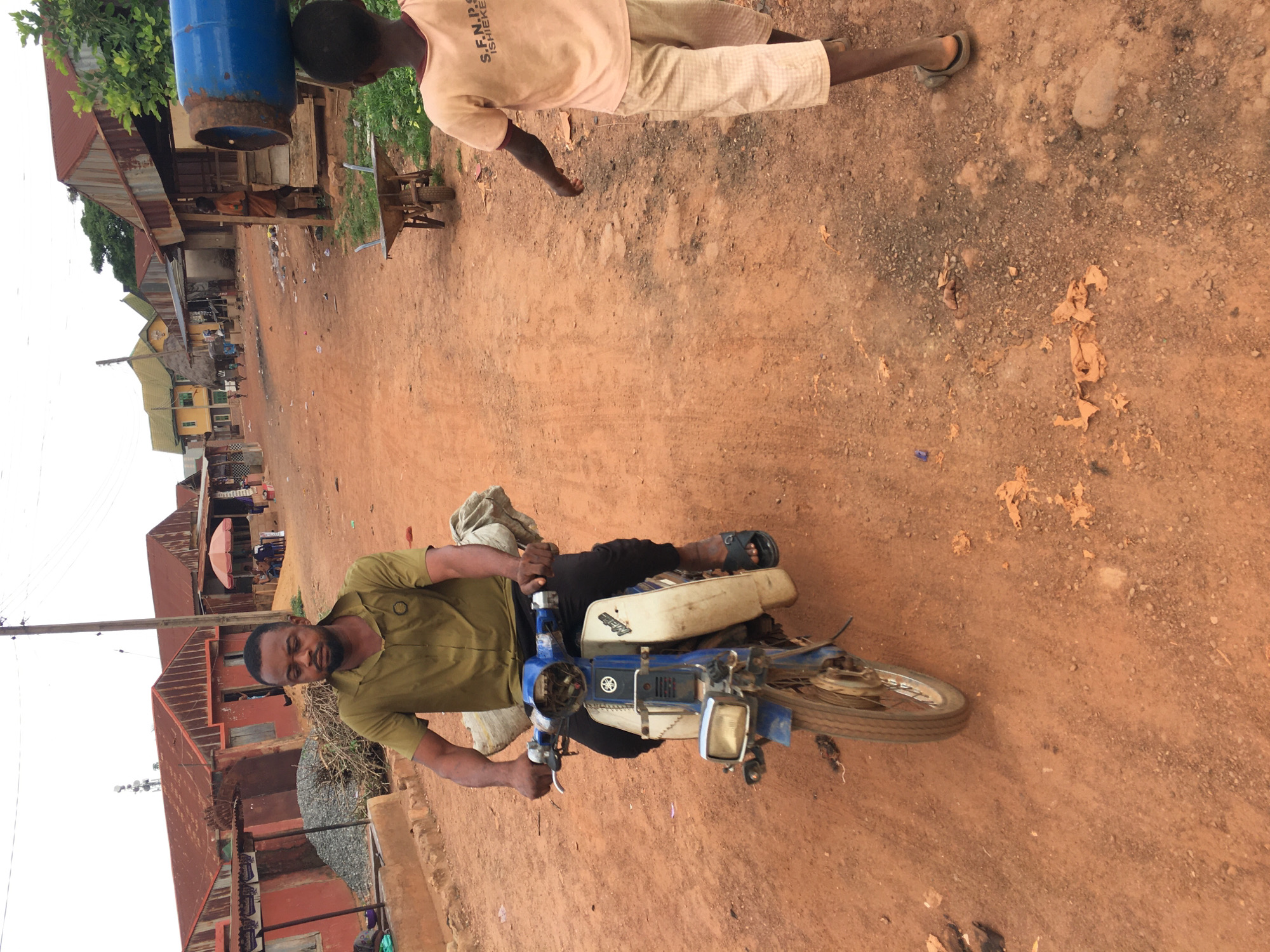 This image depicts a vibrant street scene in Isieke, Nigeria, under what appears to be an overcast daytime sky. The setting is an unpaved road characterized by reddish-brown dirt and scattered small stones, with buildings lining its edge.
In the foreground, a man is seated on a blue and white motorcycle, likely a moped or small utilitarian model, which shows signs of wear. He is dressed in a khaki-colored short-sleeved shirt and dark trousers or shorts, wearing sandals. He holds the handlebars and looks directly towards the viewer, appearing calm.
Slightly behind and to the right of the man on the motorcycle, a younger person or child stands on the dirt road, facing away from the camera. This individual is actively carrying a large, blue gas cylinder, which rests on their left shoulder. They wear a light-colored short-sleeved t-shirt with the text "S.F.N.P.O. ISIEKE" visible on the back, and light-colored checkered shorts.
The background features a row of buildings, suggesting a local community or commercial street. These structures predominantly have reddish-brown corrugated iron roofs, with walls of various colors, some painted orange or red. Sparse vegetation, including trees, and a utility pole are also visible, further defining the rural or semi-urban context. The scene captures a moment of everyday life, highlighting modes of transport and daily tasks in the area.