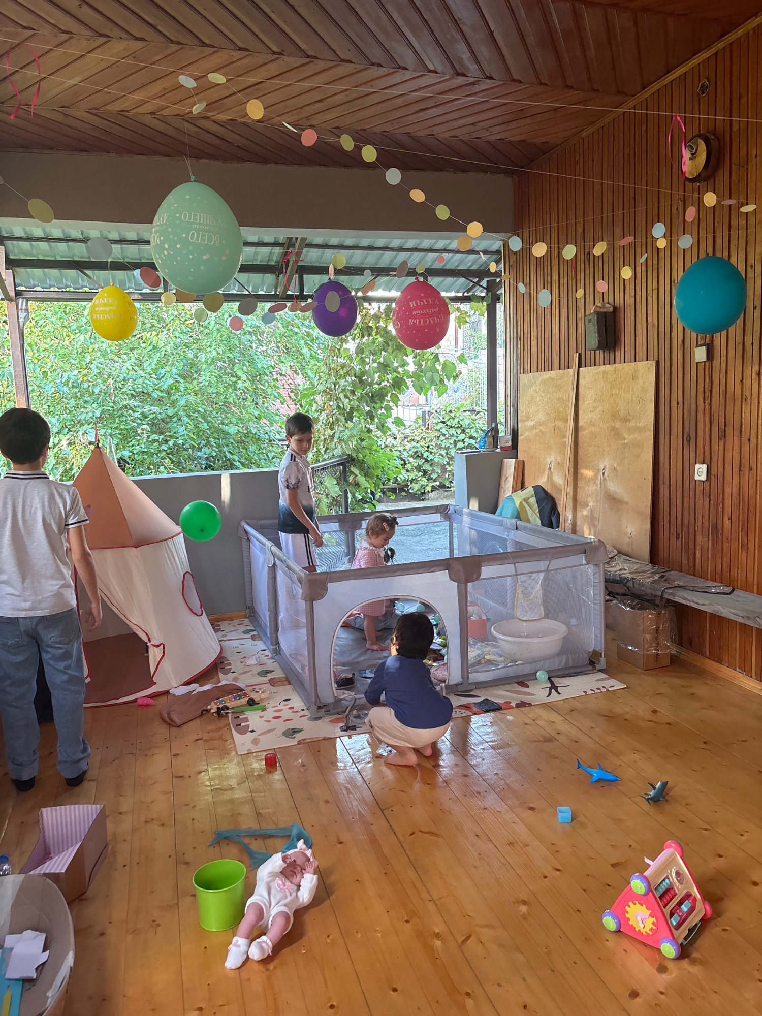 The image depicts an outdoor covered patio area with wooden floors and walls. Several children are present. In the center of the patio, there is a playpen containing a young child. A boy stands next to the playpen, looking in. Another boy is positioned on the left side of the image, facing the playpen. On the floor, a baby doll is lying down next to a green bucket. Various toys are scattered around the floor. A play tent is set up to the left of the playpen. The patio is decorated with colorful balloons and a paper circle garland. The background shows lush green foliage, suggesting an outdoor setting. The overall scene appears to be a casual gathering or playdate.