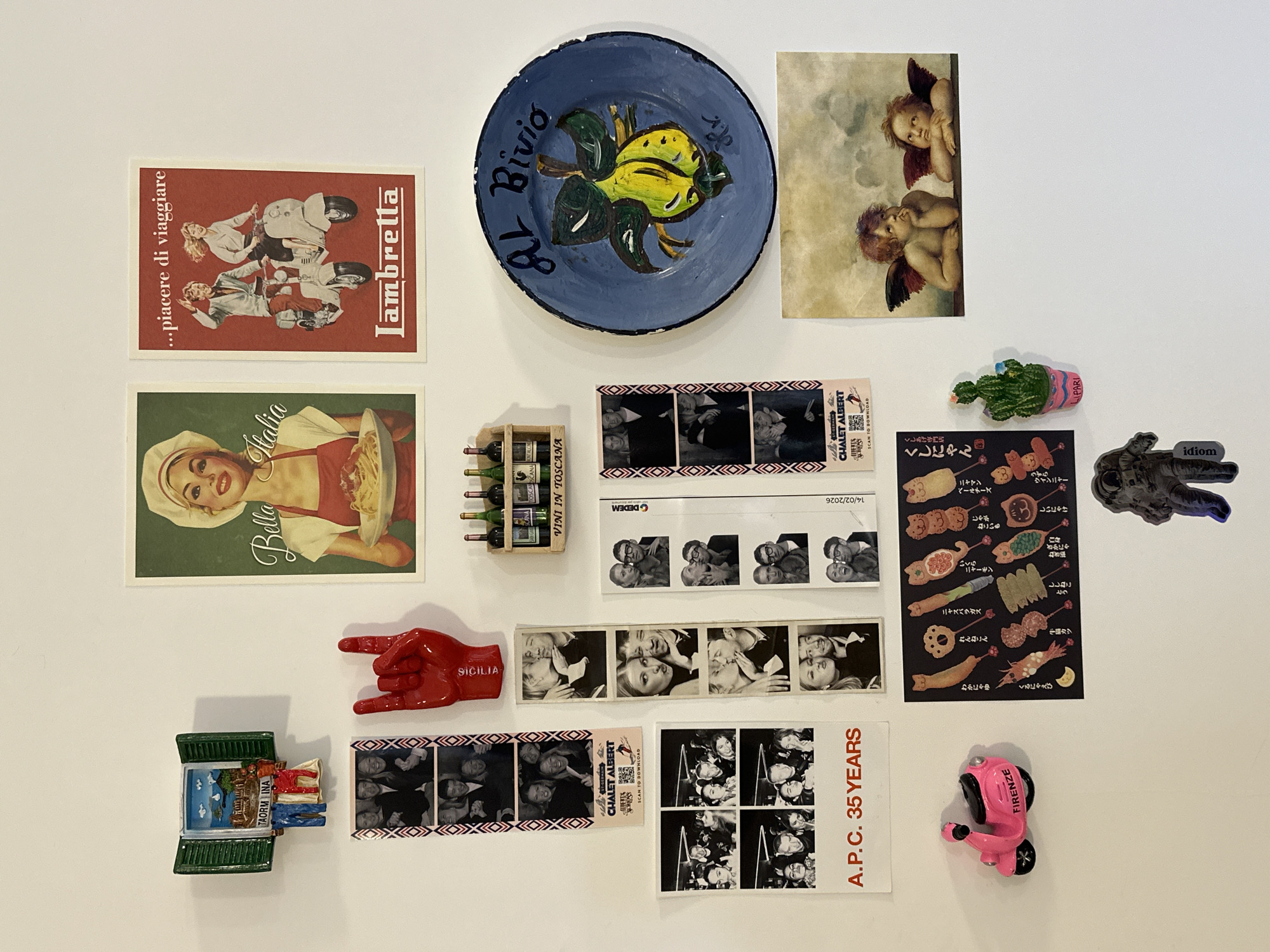 The image shows a white wall covered with various items.
Posters/Pictures:
* A red poster with the text "...piacere di viaggiare" and "Lambretta," depicting people on scooters.
* A green poster with the text "Bella Italia" and an image of a woman with a plate of pasta.
* A print of two cherubs.
* A sticker with an illustration of various types of Japanese skewers (Kushikatsu).
* A white card with the text "A.P.C. 35 YEARS" along with grayscale images of a group of people.
Objects:
* A blue ceramic plate with a lemon design and the inscription "Al Bivio."
* A miniature wooden wine rack with several bottles labeled "VINI IN TOSCANA".
* A red hand making the "rock on" gesture with the inscription "Sicilia."
* A souvenir depicting a window of Taormina.
* Several strips of black-and-white photo booth pictures.
* A cactus magnet with "LIPARI" on the pot.
* A small statue of an astronaut with the label "idiom".
* A pink scooter-shaped magnet labeled "Firenze".
* A white strip with the text "CHALET ALBERT" and a QR code for scanning.
* A white strip with the text "DEDEM" and the date "14/02/2026".
There is no indication that this image is related to London, United Kingdom. The image rather presents a collection of Italian-themed items.