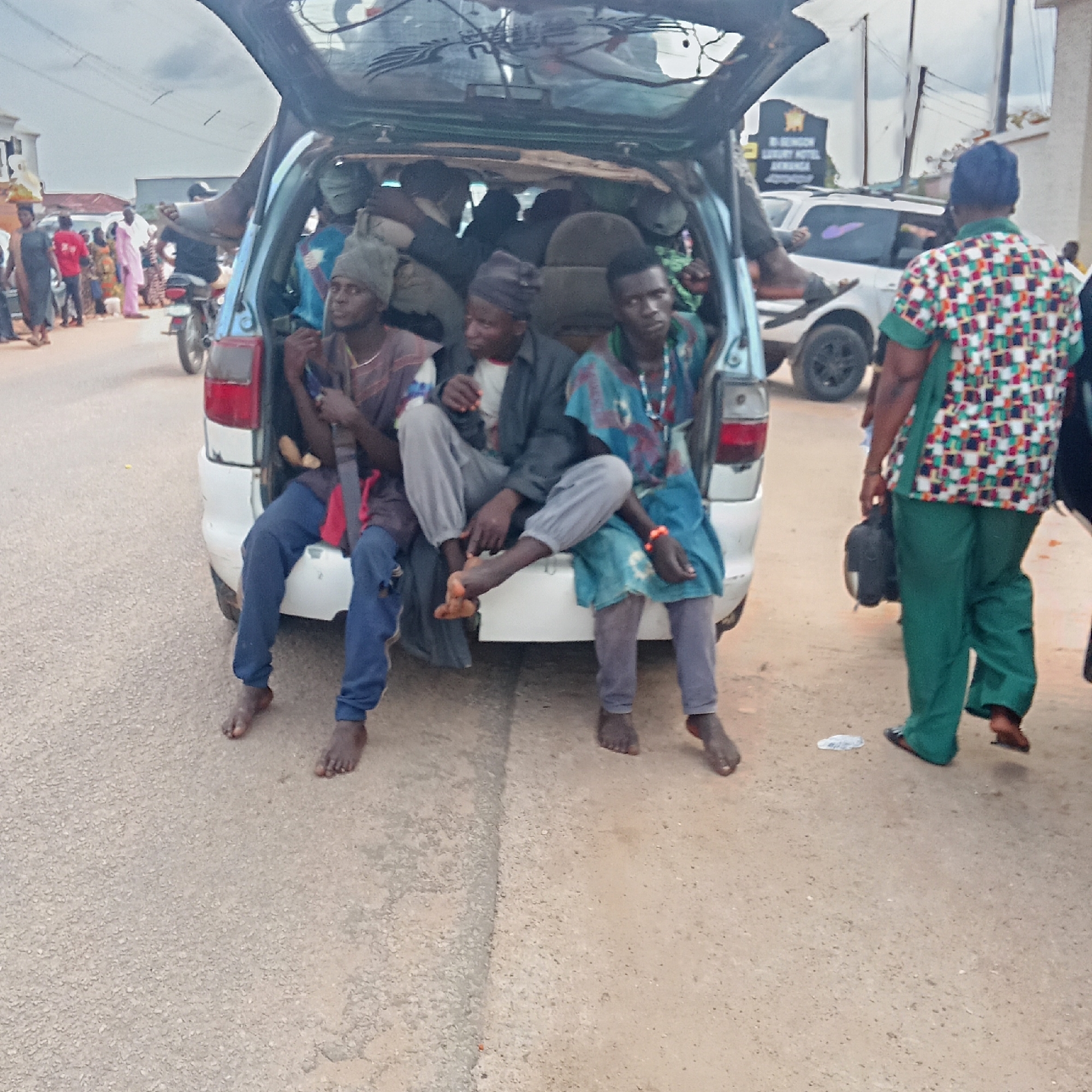 Three barefoot men are sitting in the open trunk of a light blue minivan in Akwanga, Nigeria. Additional people are inside the minivan. A man in a green patterned shirt and green pants walks to the right. People walk by on a road to the left. A white car is behind the minivan, and a sign for R. Benson Luxury Hotel Akwanga is visible in the background.