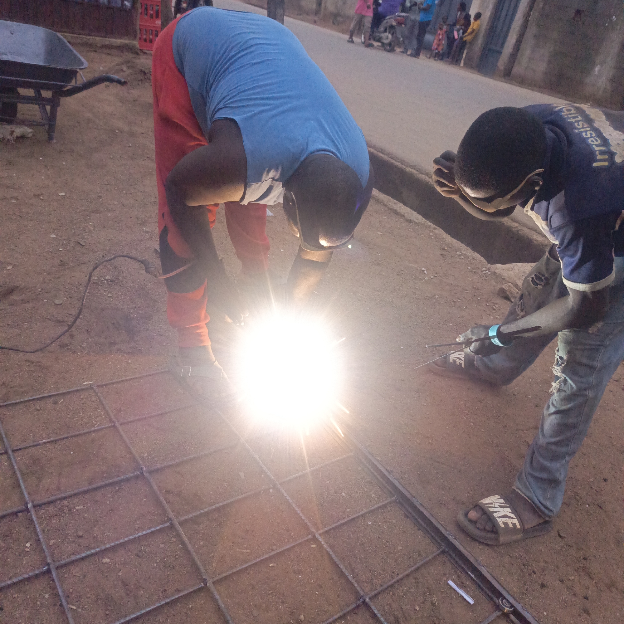 Two men are welding a metal grid on a dirt surface in Akwanga, Nigeria. One man wears orange pants, a blue shirt, and a welding mask. The second man wears ripped jeans, a dark blue shirt, and eye protection. A bright light emanates from the welding point. People are visible in the background on a road. A wheelbarrow sits to the left. The man in jeans wears Nike slides.