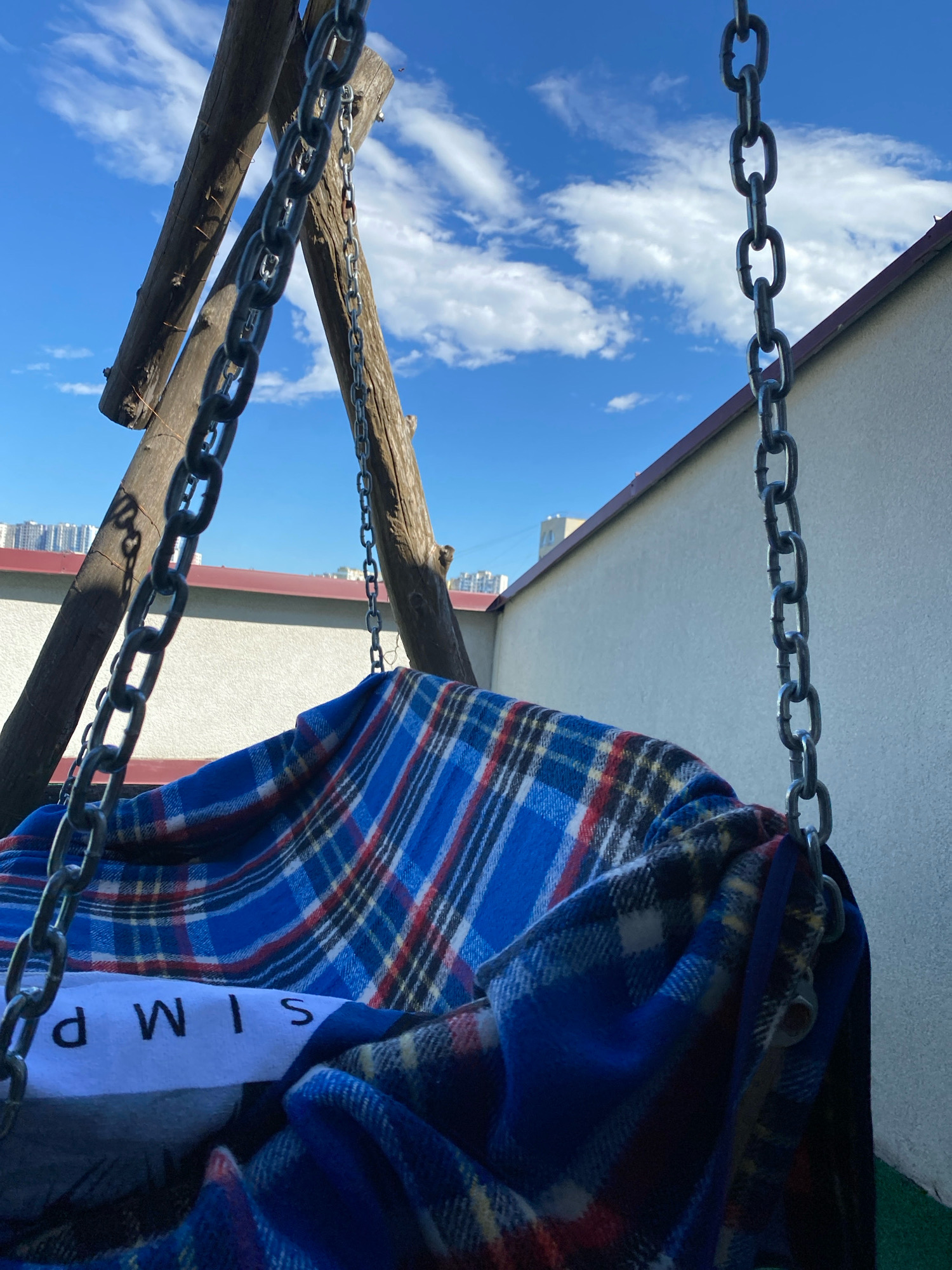 The image shows a close-up view of a wooden swing with metal chains hanging from it, partially covered by a blue, red, and white plaid blanket. A white label with black lettering, partially obscured, reads "SIM P" in reverse. The swing appears to be on a rooftop or elevated area, with a pale wall and a darker roof edge visible in the background. Above, a bright blue sky with scattered white clouds fills the upper portion of the frame. In the distance, the outlines of apartment buildings can be seen, suggesting an urban setting. The scene is well-lit, indicating it was taken during daylight hours. There are no people or animals present in the image. This scene is likely located in Chișinău, Moldova, as indicated by the context.
