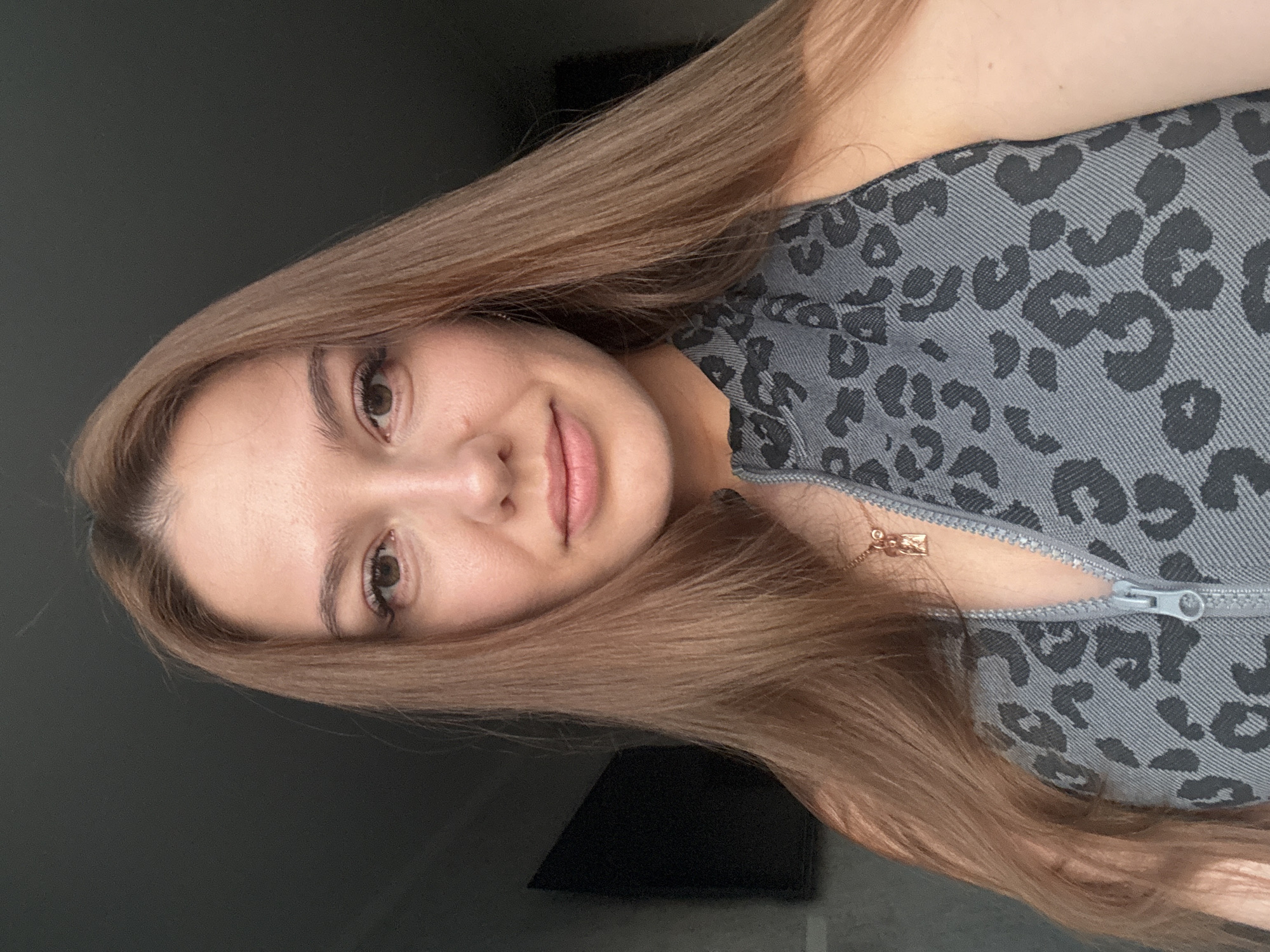 A close-up, front-facing selfie shows a young woman with long, straight, light brown hair framing her face. She is wearing a grey sleeveless top with a black leopard print and a zipper down the front, adorned with a gold pendant necklace. Her skin appears fair and smooth, with visible eyelashes and light makeup. The background is dark and appears to be an indoor setting, with a dark rectangular object, possibly a television or screen, visible on the left side. The lighting suggests a single light source from the right.