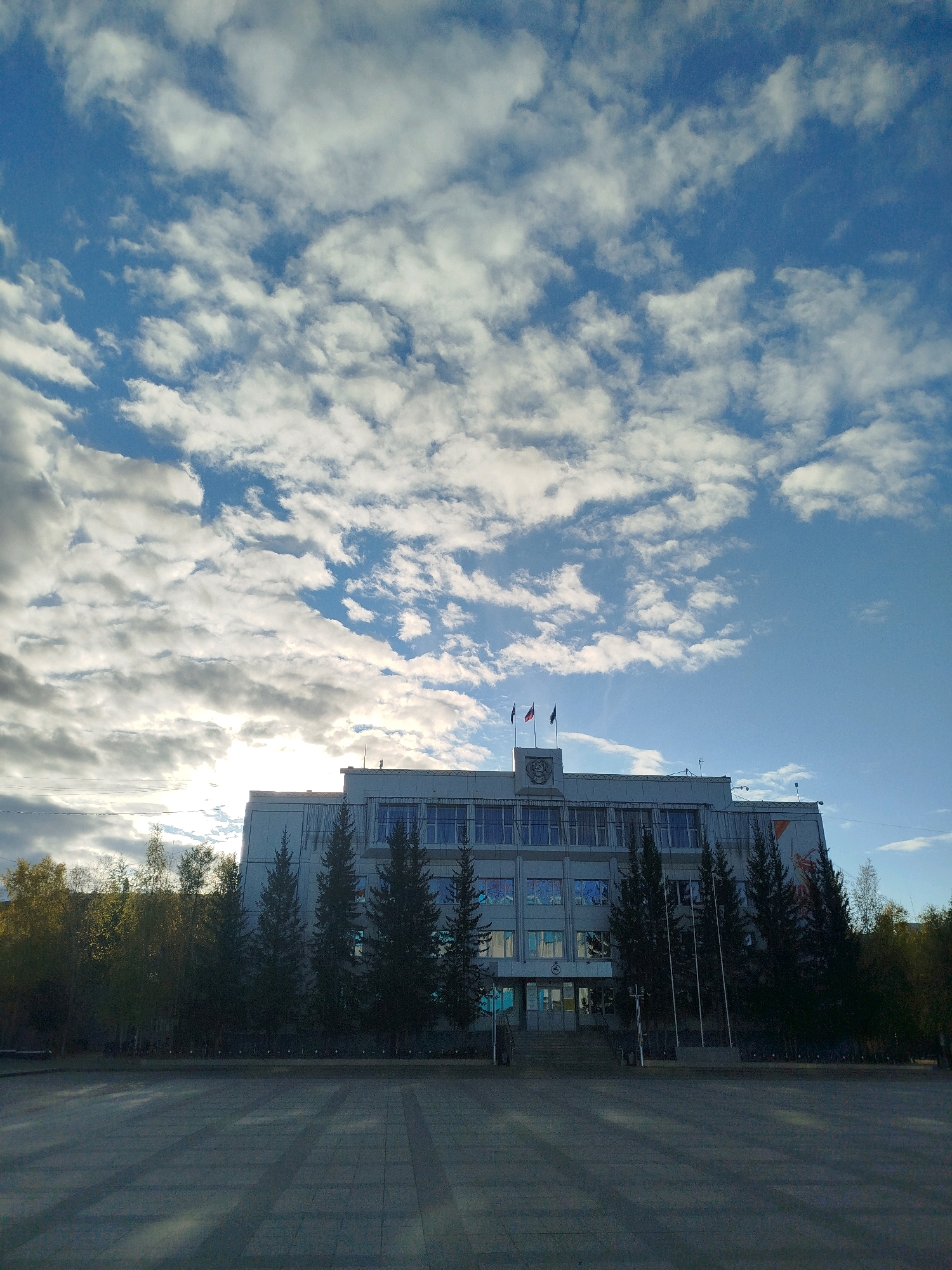 The image captures a frontal view of the Mirny City Administration building in Mirny, Russia, under a partially cloudy blue sky. The building is a multi-story, light-colored structure with numerous windows. Two flags are visible flying from the rooftop. In front of the building, there is a paved plaza and several evergreen trees are planted along the facade. The sun is low in the sky, casting long shadows across the plaza, suggesting it is either early morning or late afternoon. The surrounding area appears to be a public space, possibly a city square. There are no people or vehicles visible in the scene.