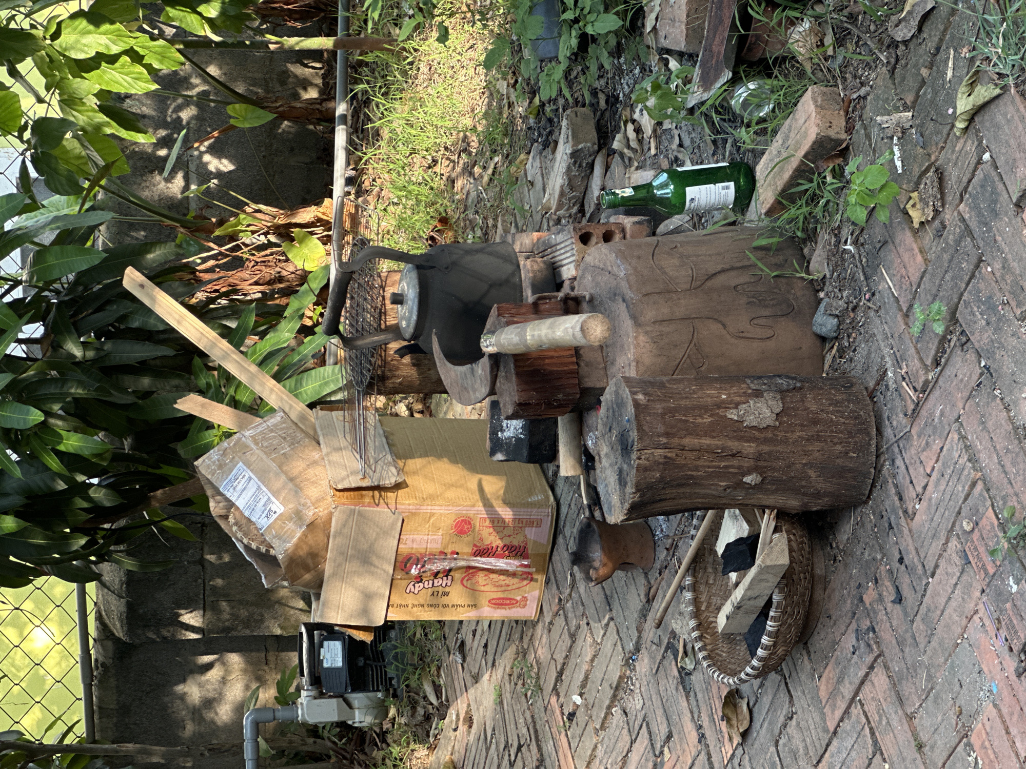 An outdoor scene in Bac Nha Trang Ward, Vietnam depicts a rustic setup, likely for cooking or a similar outdoor activity. In the foreground, a woven basket sits on a brick patio, filled with charcoal and wooden implements. Beside it, a thick, rough-hewn log serves as a base, with another, more ornate wooden object and a small, dark mortar placed nearby. A cast-iron teapot rests on a makeshift fire pit constructed from bricks, next to a collection of cooking utensils. Behind these items, an open cardboard box, partially filled with more cardboard and wooden planks, sits near a water pump and motor. Lush green foliage and trees form a natural backdrop, with a concrete wall and chain-link fence visible in the distance. The ground is paved with reddish-brown bricks, with patches of grass and scattered debris, suggesting an outdoor, potentially utilitarian space. A green glass bottle stands upright amongst the grass. The lighting indicates it is daytime, with dappled sunlight filtering through the leaves.