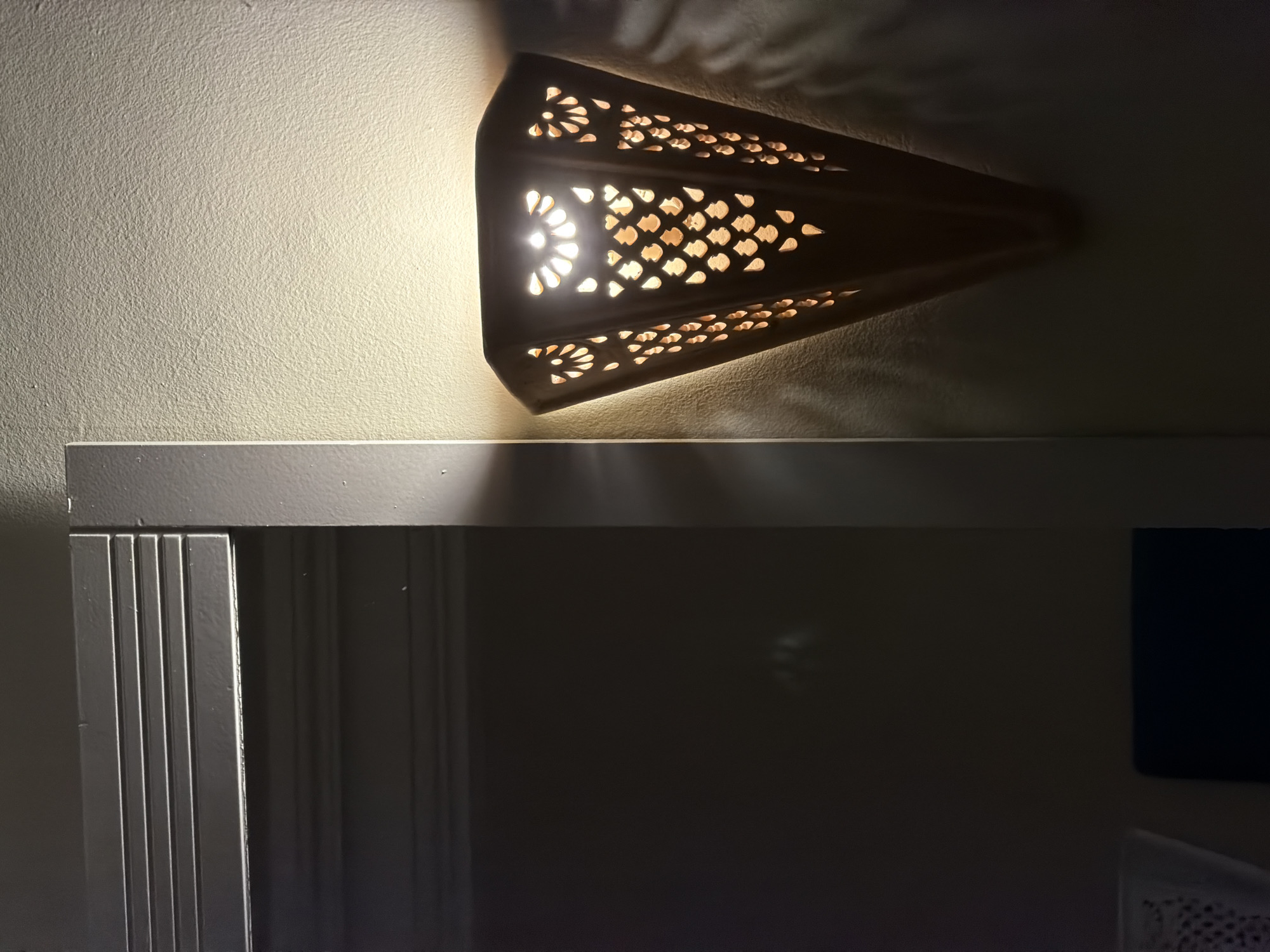 The image shows a wall-mounted lamp with an intricate cutout design, casting a warm, patterned light onto the textured cream-colored wall. The lamp itself is a dark, conical shape, wider at the top and tapering to a point. The light source within illuminates the cutouts, which resemble stylized floral or teardrop shapes, creating a dappled effect on the wall. To the left of the lamp, a portion of a dark, recessed area is visible, featuring horizontal ridged detailing on its surface, suggesting a doorway or cabinet. The overall scene is dimly lit, with the lamp being the primary light source, creating a contrast between illuminated and shadowed areas. There are no people or animals visible in the image. The setting appears to be an indoor space, possibly a hallway or a room in a home. Without further context, the specific location within the city of Sousse, Tunisia cannot be determined from the visual information alone.