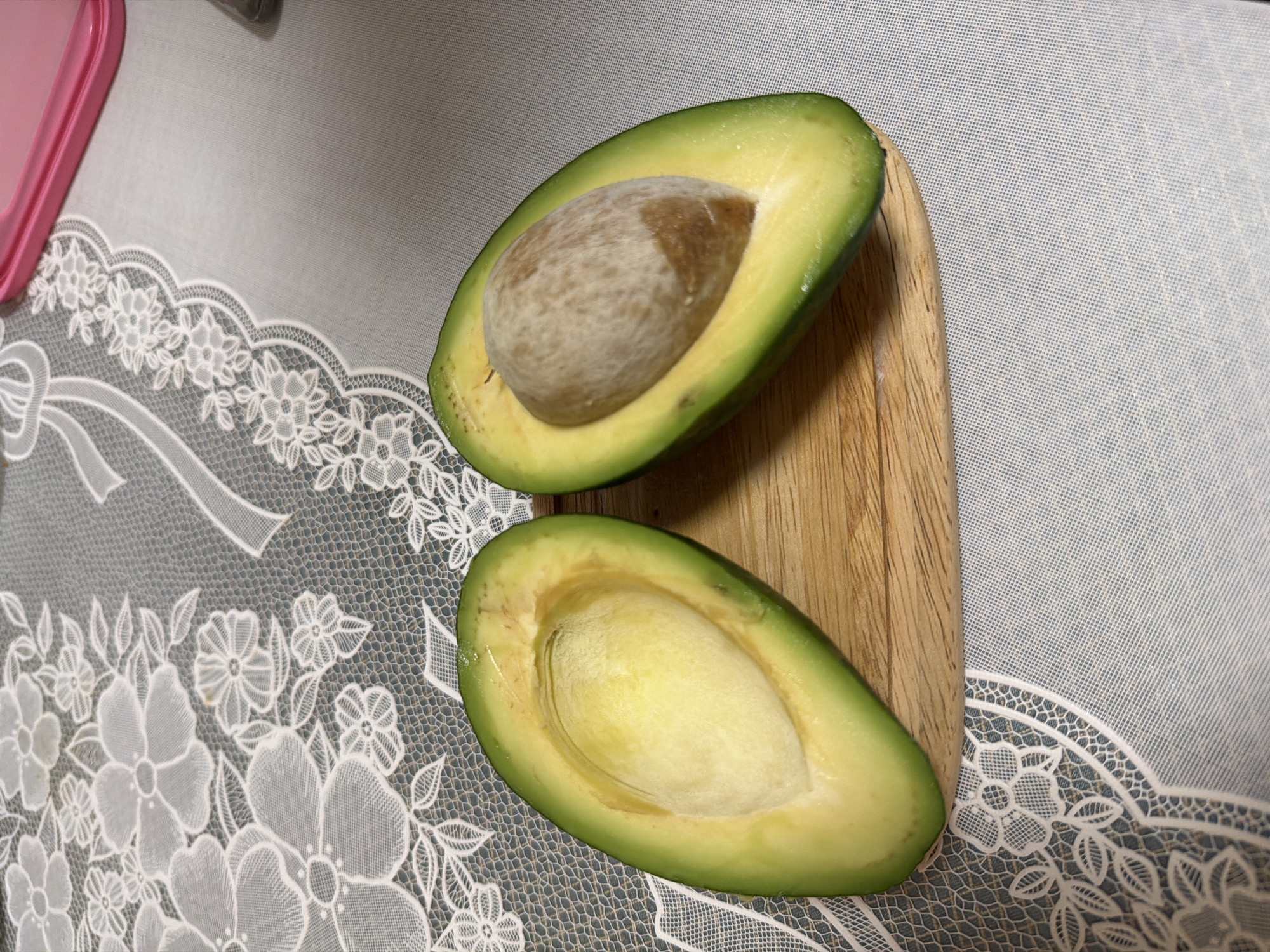 Two halves of a ripe avocado are displayed on a small wooden cutting board. The avocado on the left is hollowed out, revealing its bright yellow flesh. The avocado on the right contains a large, brown pit. The cutting board rests on a white lace tablecloth, which is placed over a grey textured surface. A pink object is visible in the top right corner of the image. The scene appears to be set indoors. There are no people present in the image.