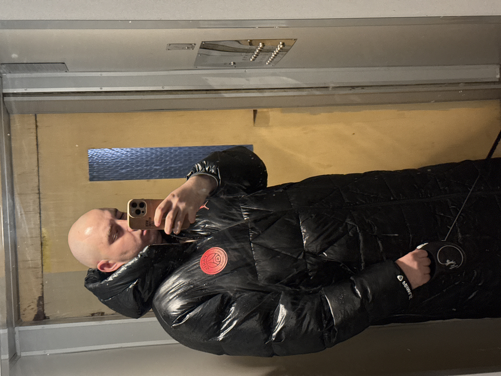 A bald man is taking a selfie in an elevator with a gold iPhone. He is wearing a black puffer jacket with a red circular patch on the chest. He is holding a black retractable dog leash in his left hand. The elevator has a wooden paneled wall with a horizontal blue window near the top. Above the door, there is a panel with multiple elevator buttons. The location cannot be determined as Pleven, Bulgaria, from the visual information provided.