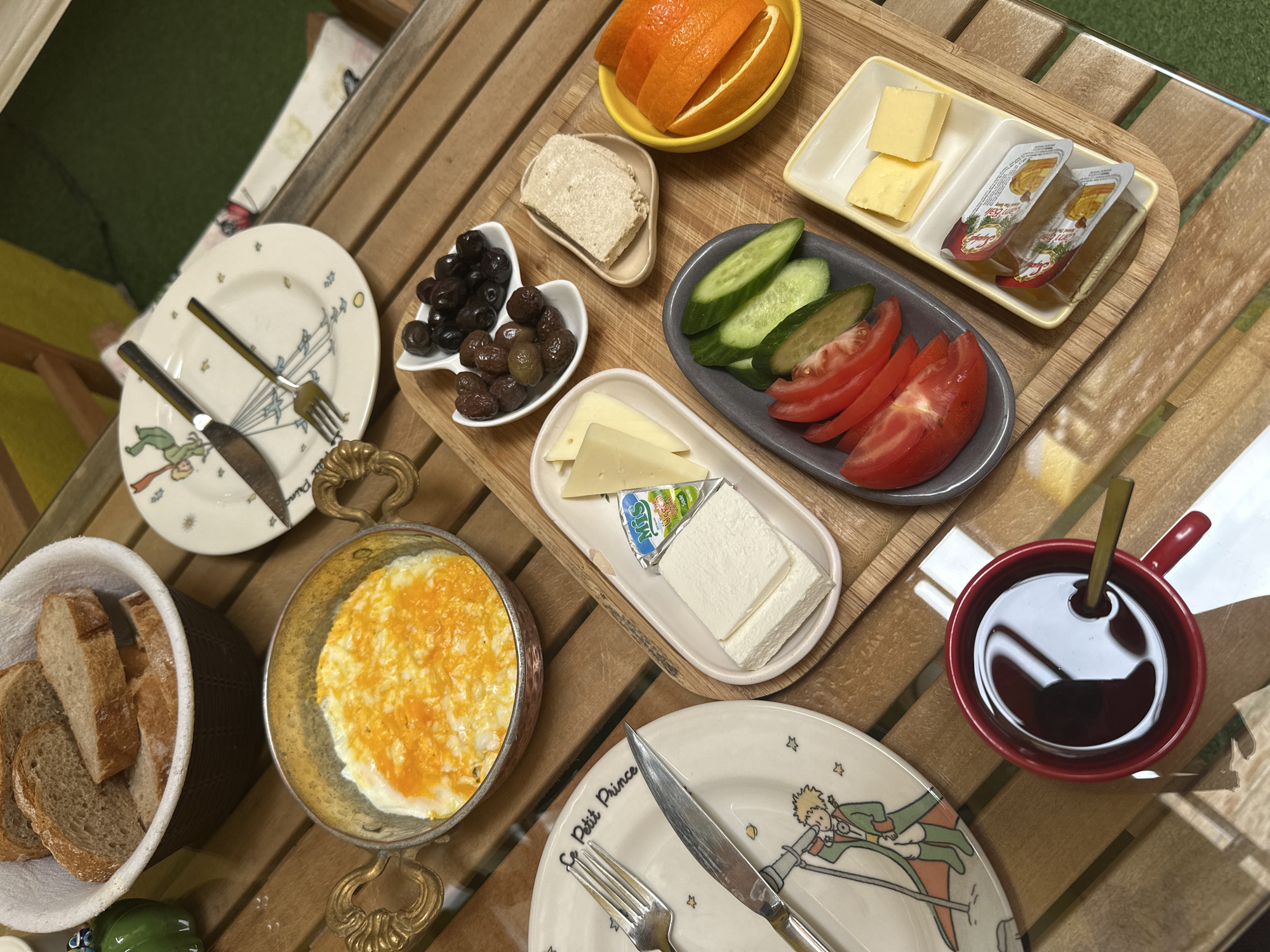The image shows a breakfast spread on a wooden table in Antalya, Turkey. There is a variety of food items including sliced oranges, cheese, jam, cucumber, tomatoes, olives, tahini, bread, menemen (Turkish egg dish), and a cup of black coffee. There are plates with "Le Petit Prince" (The Little Prince) design, cutlery, and a bread basket. The table is made of light wood and there is a glimpse of greenery in the background.