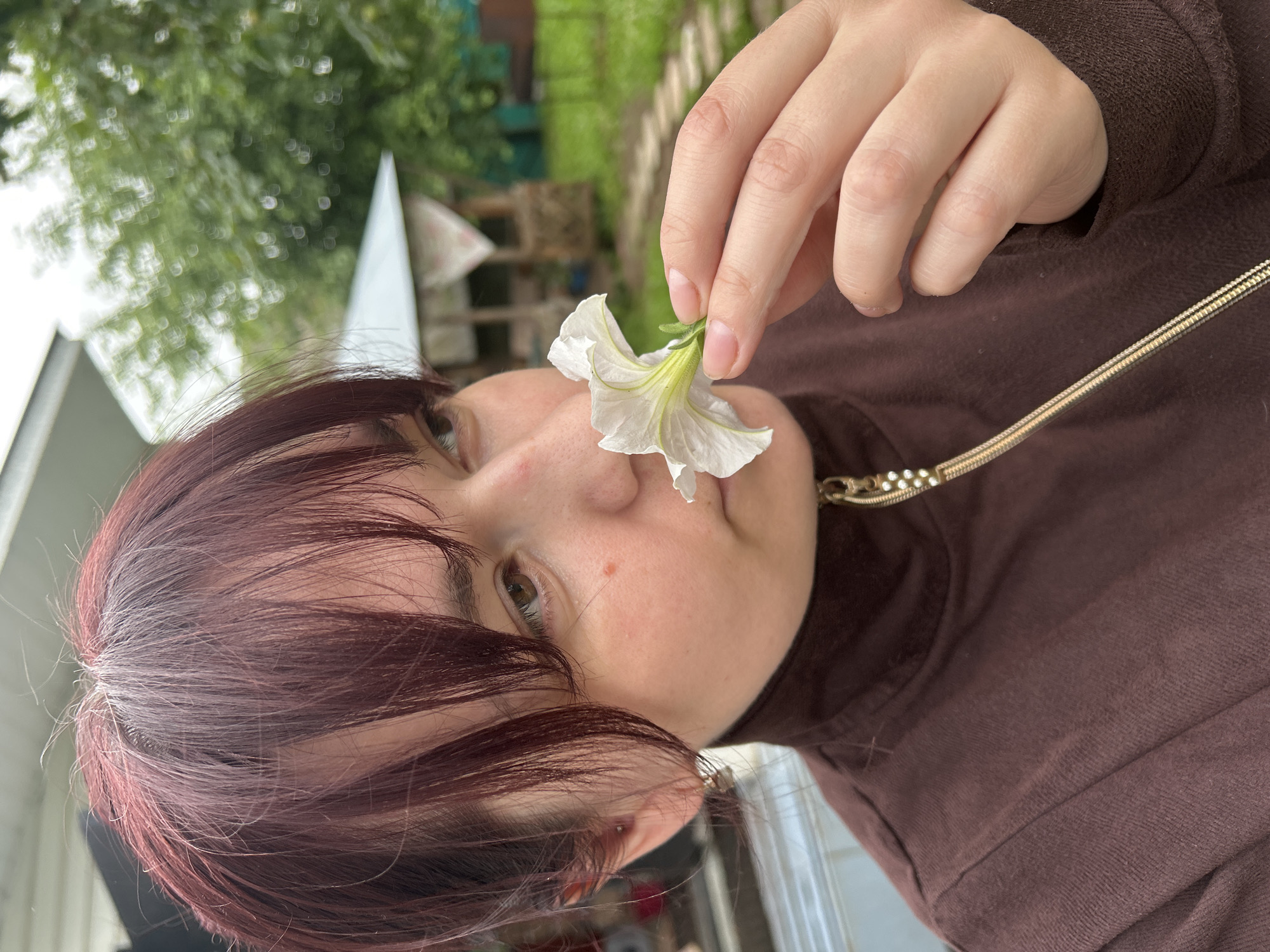 A young person with dark, reddish-brown hair is captured in a close-up shot, holding a delicate white flower near their nose. The flower, with its pale petals and greenish-yellow center, is delicately positioned, suggesting a moment of gentle interaction with nature. The person's face is partially obscured by their hair, but their right eye is visible, looking slightly off-camera. A small red mark is visible on their cheek. They are wearing a dark brown turtleneck sweater, adorned with a gold chain necklace that is partially visible.
The background is softly blurred, indicating a shallow depth of field. Hints of green foliage and some indistinct structures suggest an outdoor setting, possibly a garden or a backyard, consistent with a location context in Фоки, Russia. The lighting appears to be natural, suggesting daytime, and the overall mood is serene and contemplative. The image is oriented vertically. No text is visible in the frame.
