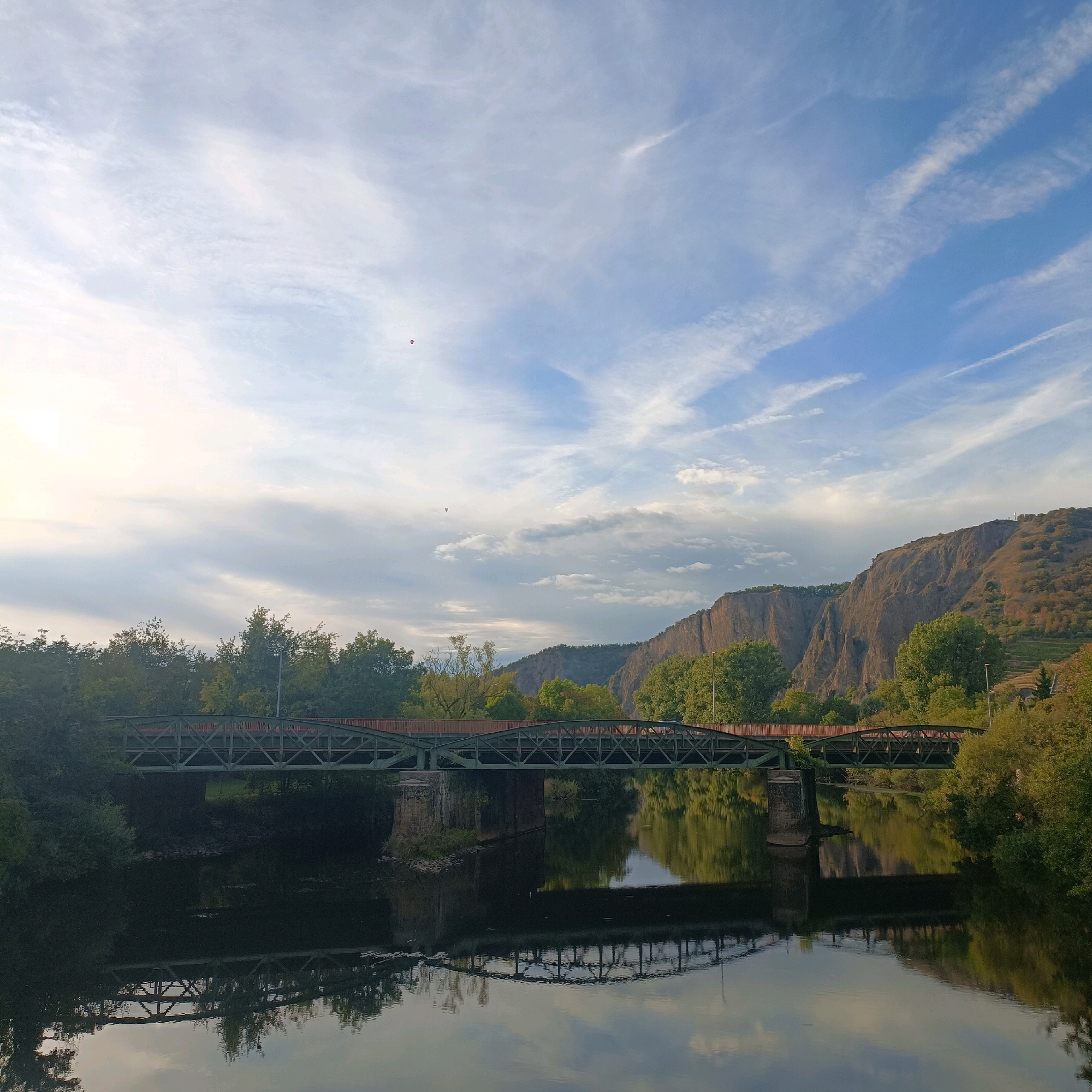 The image depicts a scenic view of the Nahe River in Bad Kreuznach, Germany, at sunrise. A historic railway bridge, characterized by its green metal structure and intricate latticework, spans the river. The bridge is reflected in the calm water, creating a symmetrical composition. Lush green trees line the riverbanks and the steep, rocky slopes of the Klause, a prominent landmark in the city. The sky is adorned with soft, wispy clouds, painted with the gentle hues of the rising sun. Two hot air balloons are visible in the distance, adding a touch of vibrancy to the tranquil scene. The overall atmosphere is serene and picturesque.