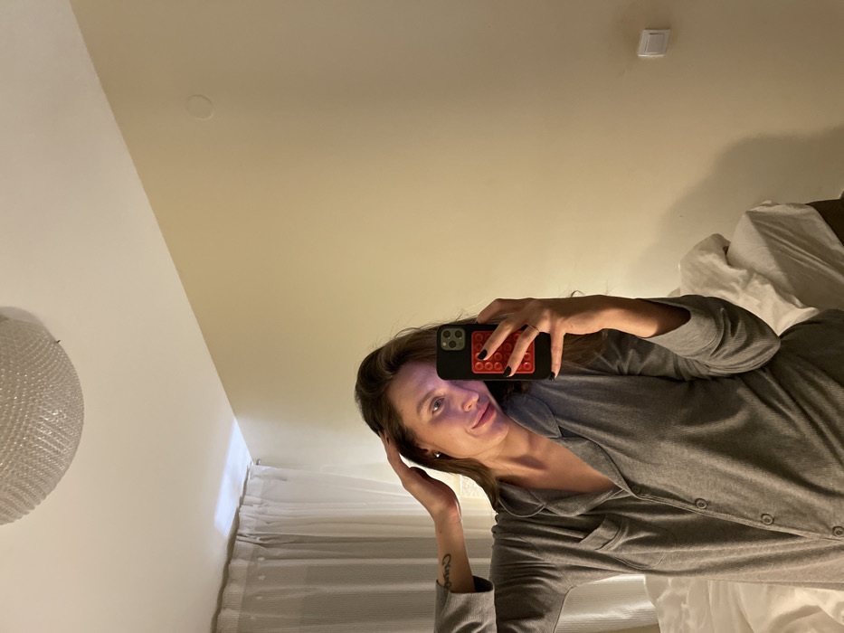A woman in gray pajamas poses for a selfie. She holds her phone, which has a colorful case, with her right hand, partially obscuring her face. Her left hand touches her hair. She lies in a bed with white sheets and a white headboard. A patterned white light fixture is on the left wall. A light switch is visible near the ceiling. She has a tattoo on her left forearm.