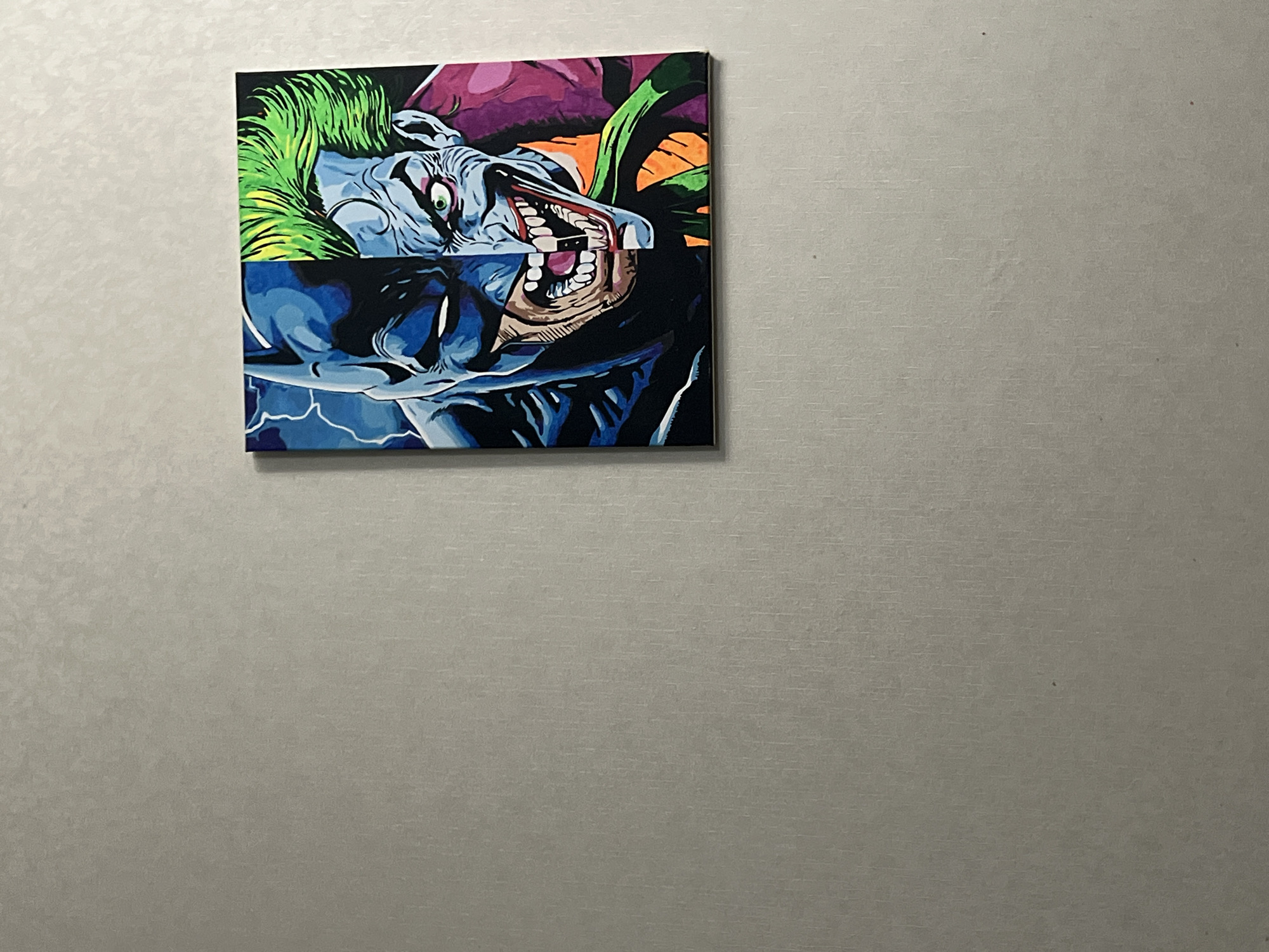 A square artwork hangs on a neutral-colored wall. The artwork appears to be a stylized, comic-book-inspired depiction of Batman and the Joker, split horizontally. The upper half shows the Joker with green hair, a pale face, and a menacing grin, while the lower half displays Batman in a dark blue suit with lightning-like energy emanating from his body. The image is of a wall in Chaikovsky, Russia.