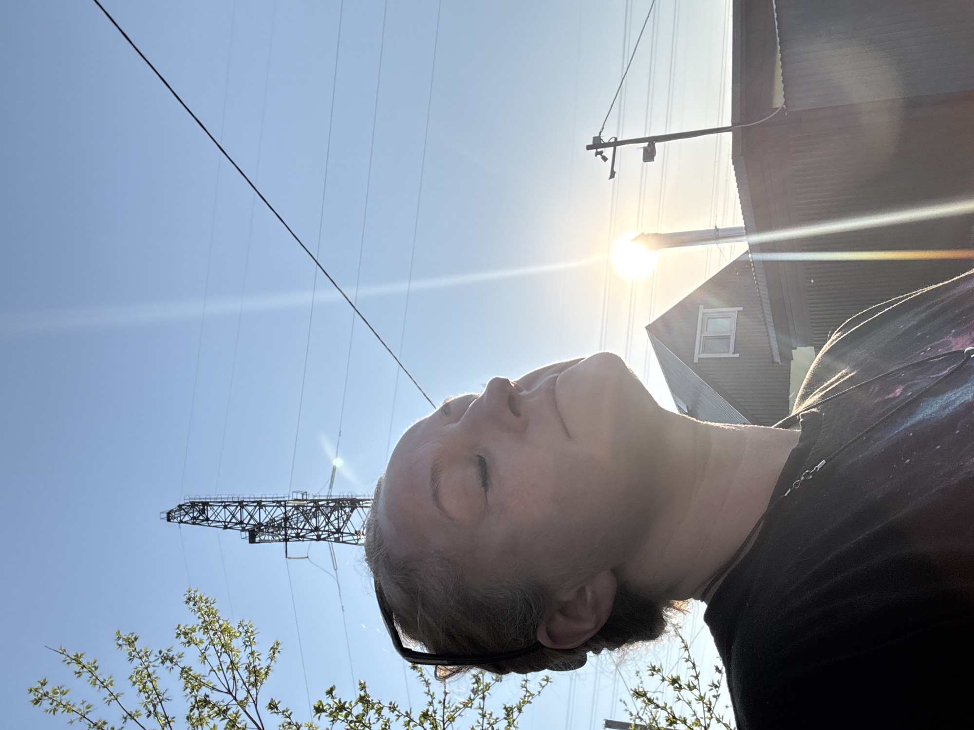 A low-angle shot of a person's face, with eyes closed, facing the sun. The background shows a clear, bright blue sky with overhead power lines and an electrical transmission tower on the left. To the right, a building with a dormer window is partially visible. The scene is lit by the sun, with lens flare. The lower left features blossoming tree branches with new green leaves. A necklace hangs from a black t-shirt. The specific location in the city of Chaikovsky, Russia, cannot be identified from this image.