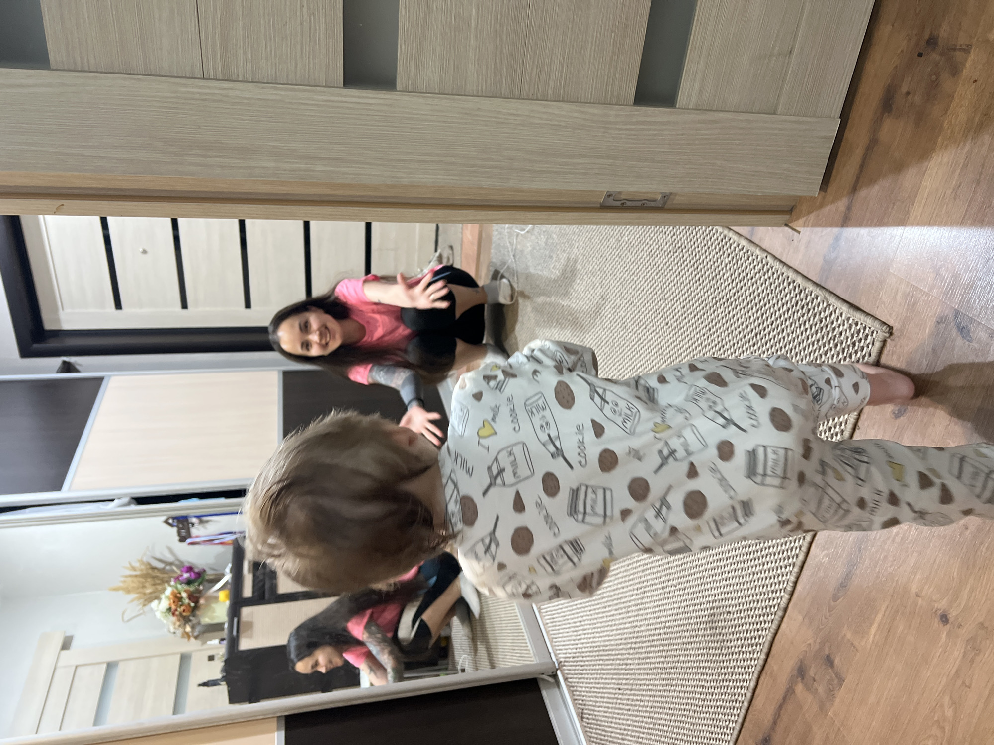 This image, captured in the city of Noviy, Russia, depicts an indoor scene with a young child and an adult woman interacting. In the foreground, a toddler, viewed from behind, stands on a beige textured rug. The child wears a white sleepsuit patterned with brown cookies and white milk cartons/bottles, some bearing "MILK" text and yellow hearts. Small pink foot coverings are visible. The child's light brown hair is slightly disheveled.
In the mid-ground, a young adult woman crouches on the rug, facing the child, with her arms extended forward and palms open in an inviting gesture. She wears a bright pink t-shirt and dark pants, with white socks visible on her feet. Tattoos are visible on her left arm. The woman is smiling broadly, directly engaging the child.
A large, light-framed mirror on the left side of the scene reflects the woman and additional elements of the room. The reflection shows a dark brown and beige cabinet behind the woman, adorned with a vase holding dried wheat-like stalks and fresh colorful flowers (orange, white, purple).
To the right of the woman, a light-colored wooden interior door with horizontal panels is partially open, revealing a recessed latch mechanism. Further in the background, beyond the woman, a different light-colored door, possibly an entryway door, features five horizontal dark stripes and a metallic peephole. This door is framed by a darker border. The floor transitions from the beige rug to a light brown wooden surface on the left, right, and foreground.
