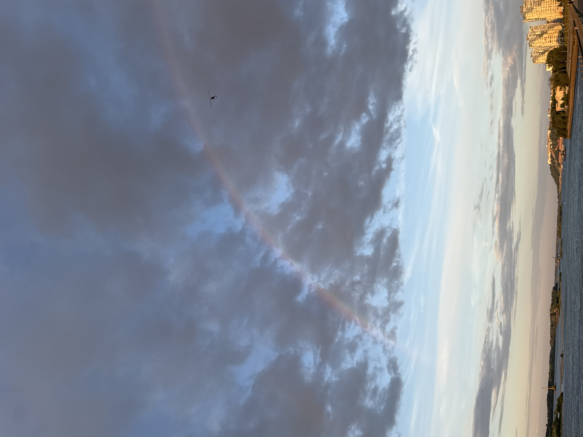 This media file captures a sky view with a faint rainbow arching through dark, dramatic clouds on the left. A silhouette of a helicopter is visible against the darker clouds. On the right, the scene transitions to a brighter, clearer sky with lighter clouds, revealing a cityscape with tall buildings and trees lining a waterfront. The calm water of a river or bay occupies the bottom right of the image. The juxtaposition of the stormy sky with the serene cityscape suggests a fleeting moment of a rainbow after or during changing weather. The buildings and waterfront are bathed in the warm, soft light of either sunrise or sunset, indicating early morning or late evening. The overall mood is one of natural beauty and urban tranquility. There is no visible text in the image.