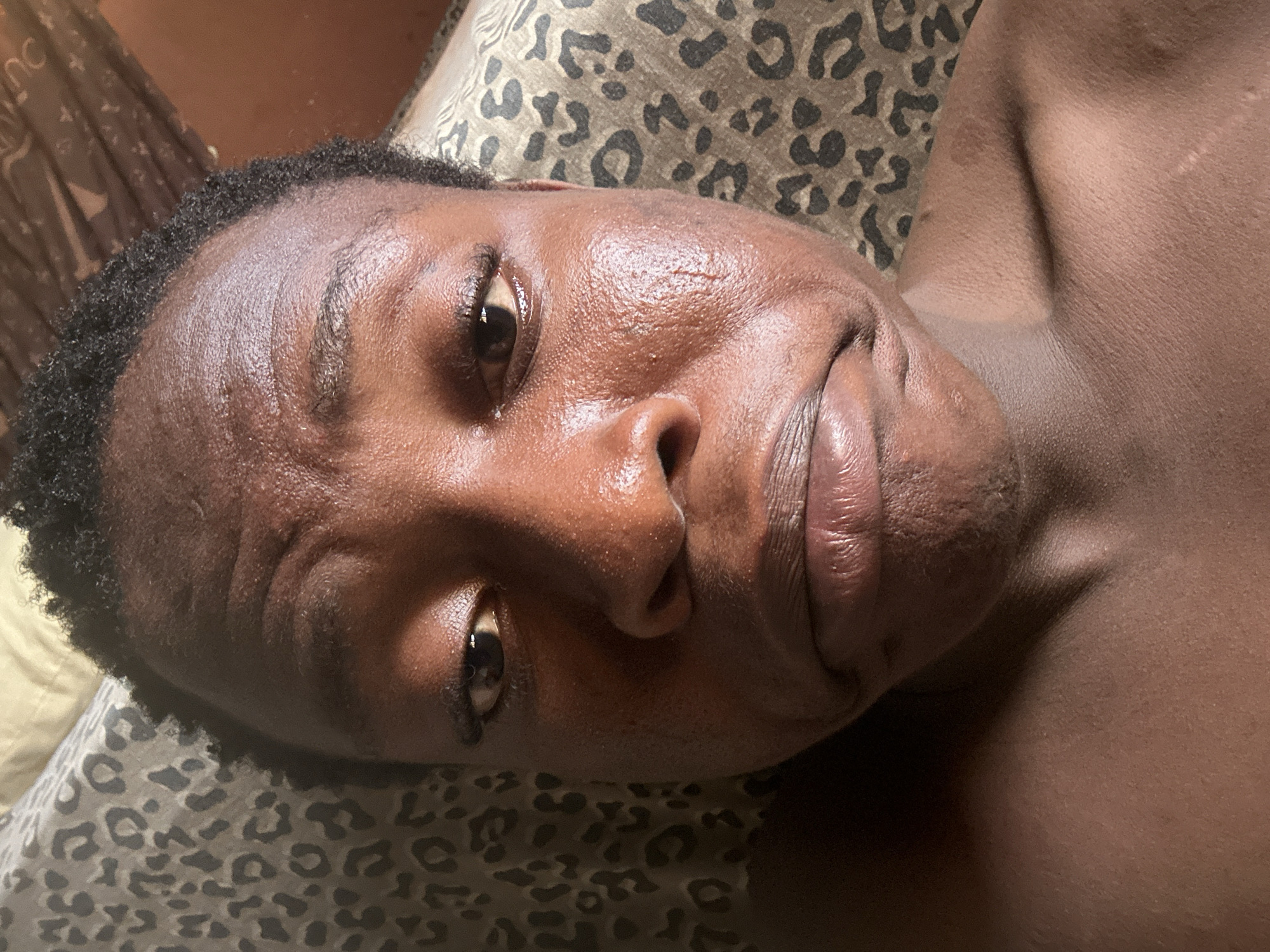 A close-up, high-angle shot depicts a person with dark skin lying on what appears to be a bed with a leopard-print cover. The person's face is the primary focus, showing details such as small wrinkles on the forehead, distinct facial features, and reflective light on the skin. The background is slightly blurred, with a glimpse of other bedding or furniture. There is no indication of a location in the city of Unknown, Unknown, or any specific actions or interactions beyond the person simply lying down. The environment is limited to the immediate surroundings of the bedding and person.