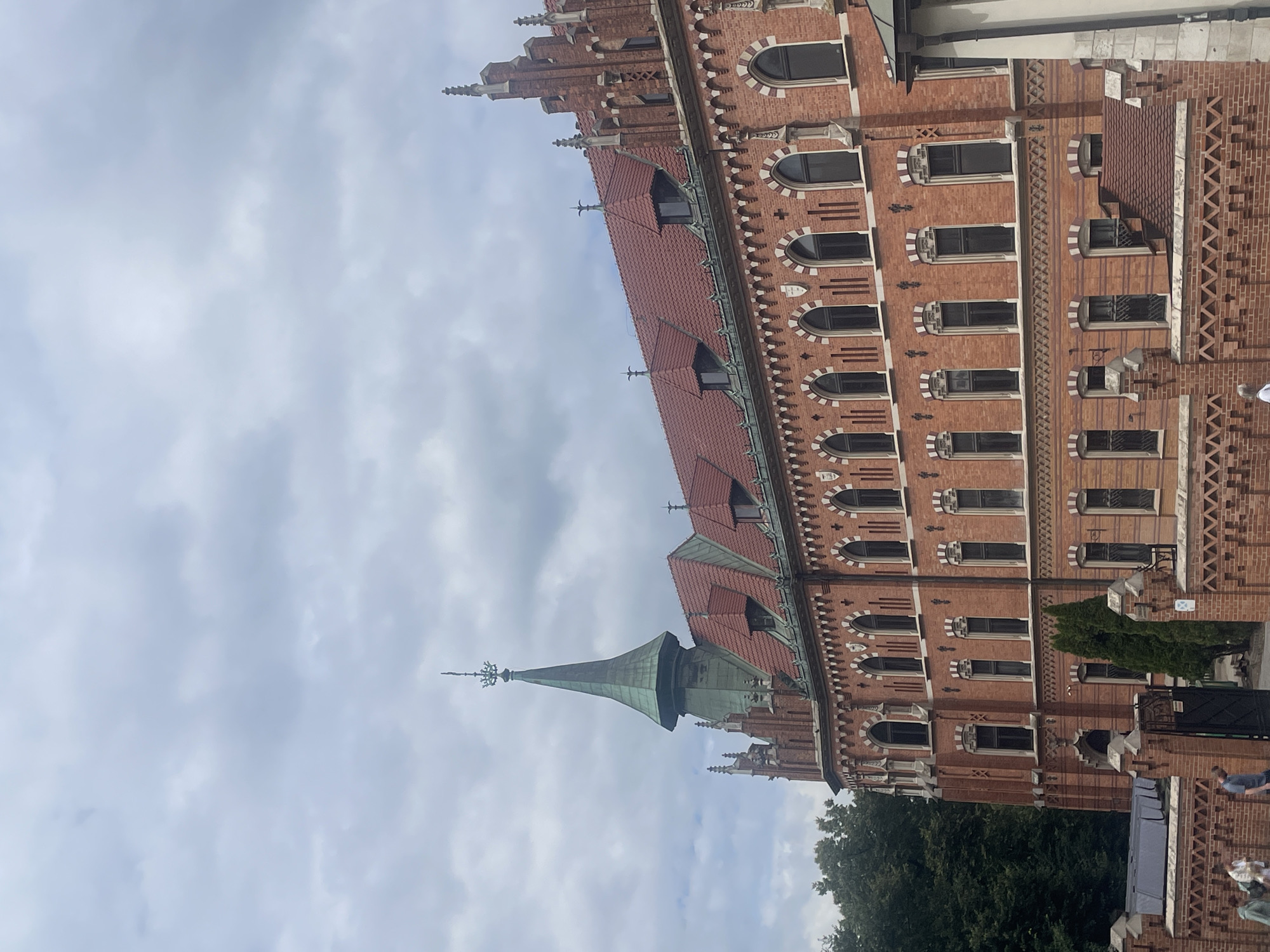 This is a medium-shot, exterior photograph of the Collegium Minus building of Jagiellonian University in Krakow, Poland. The building is constructed of red brick with a red tile roof. It has a tall, green, conical spire on the left side, and several smaller spires and decorative elements on the roofline. The windows are arched, and the building features intricate brickwork detailing. In the foreground, there is a low brick wall with a metal gate. Several people are visible walking along the pavement behind the wall. The sky is overcast and cloudy. Trees are visible to the left of the building.